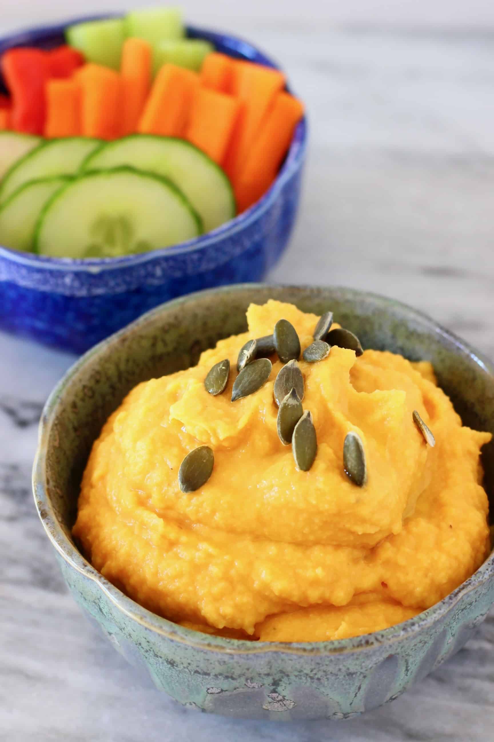 A grey bowl of orange pumpkin hummus topped with green pumpkin seeds and a blue bowl of chopped vegetables on a marble surface