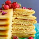 A stack of cornmeal pancakes with a slice cut out of them topped with rose petals and small strawberries on a green plate against a grey background