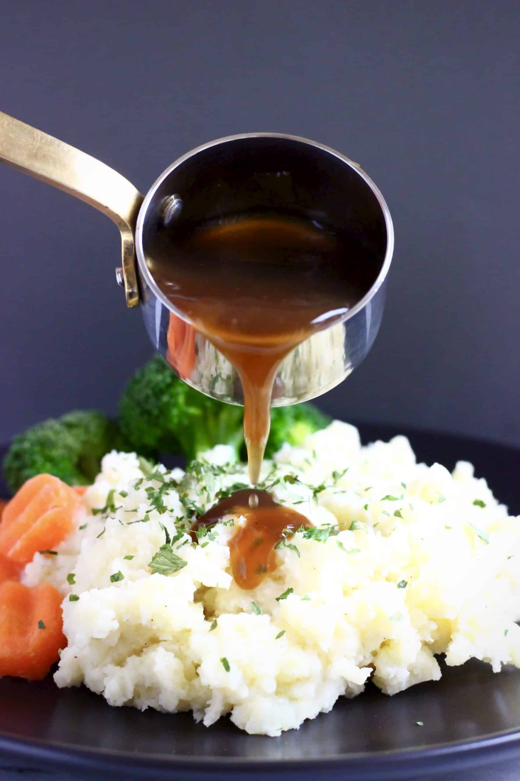 Mashed potatoes topped with green herbs with sliced carrots and broccoli on a black plate with brown gravy being poured over
