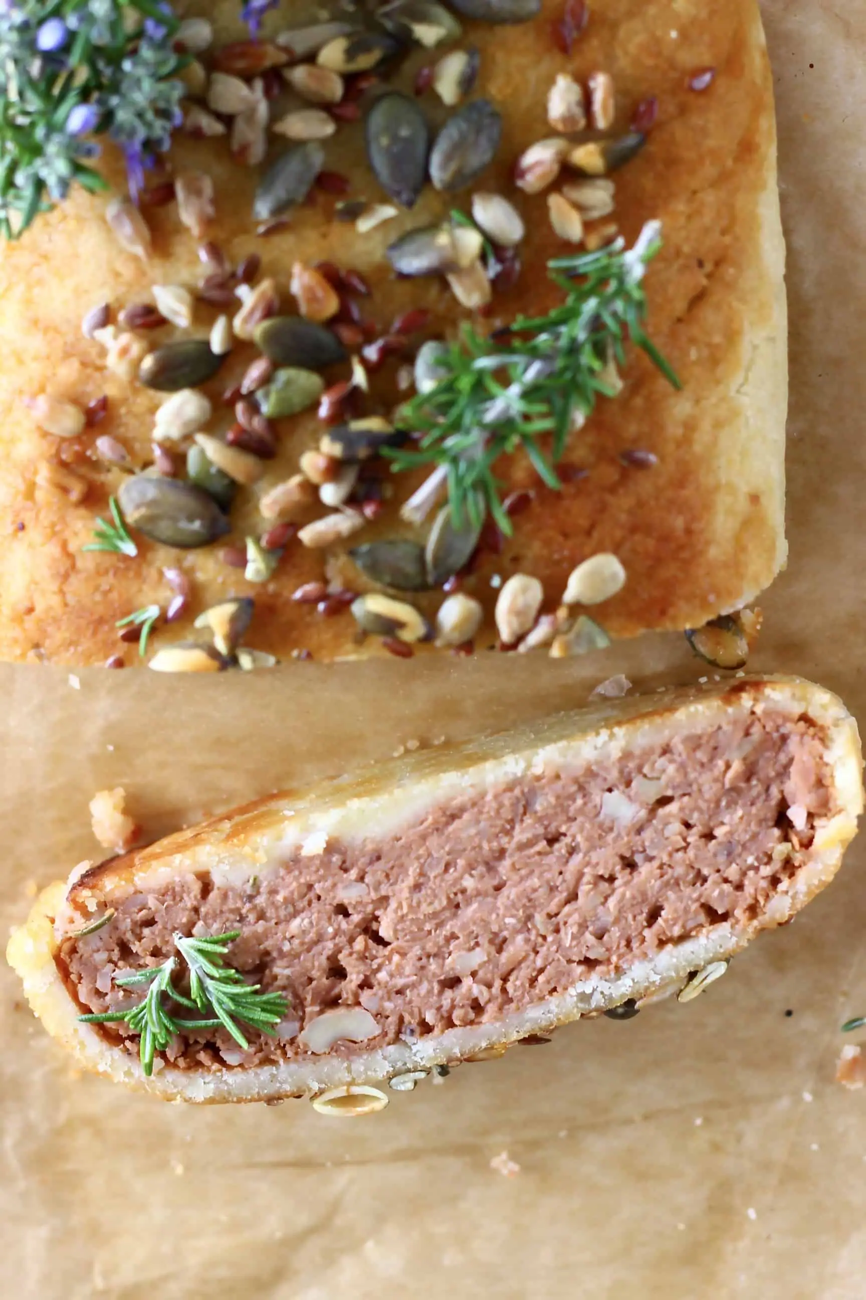 A vegan Wellington topped with mixed seeds with a slice of it on the side on a sheet of brown baking paper