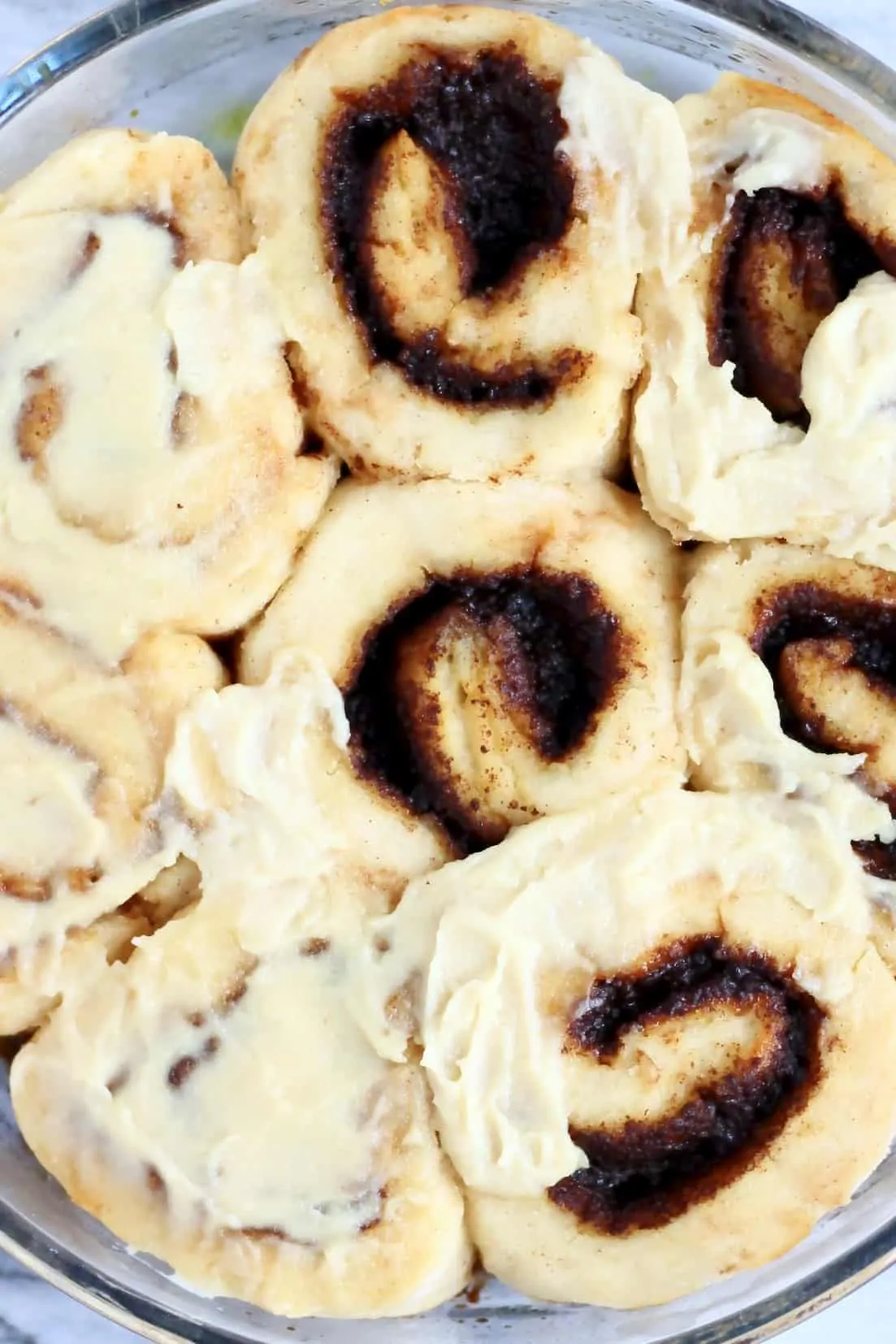 Seven gluten-free vegan cinnamon rolls in a round baking dish, topped with vegan cream cheese frosting