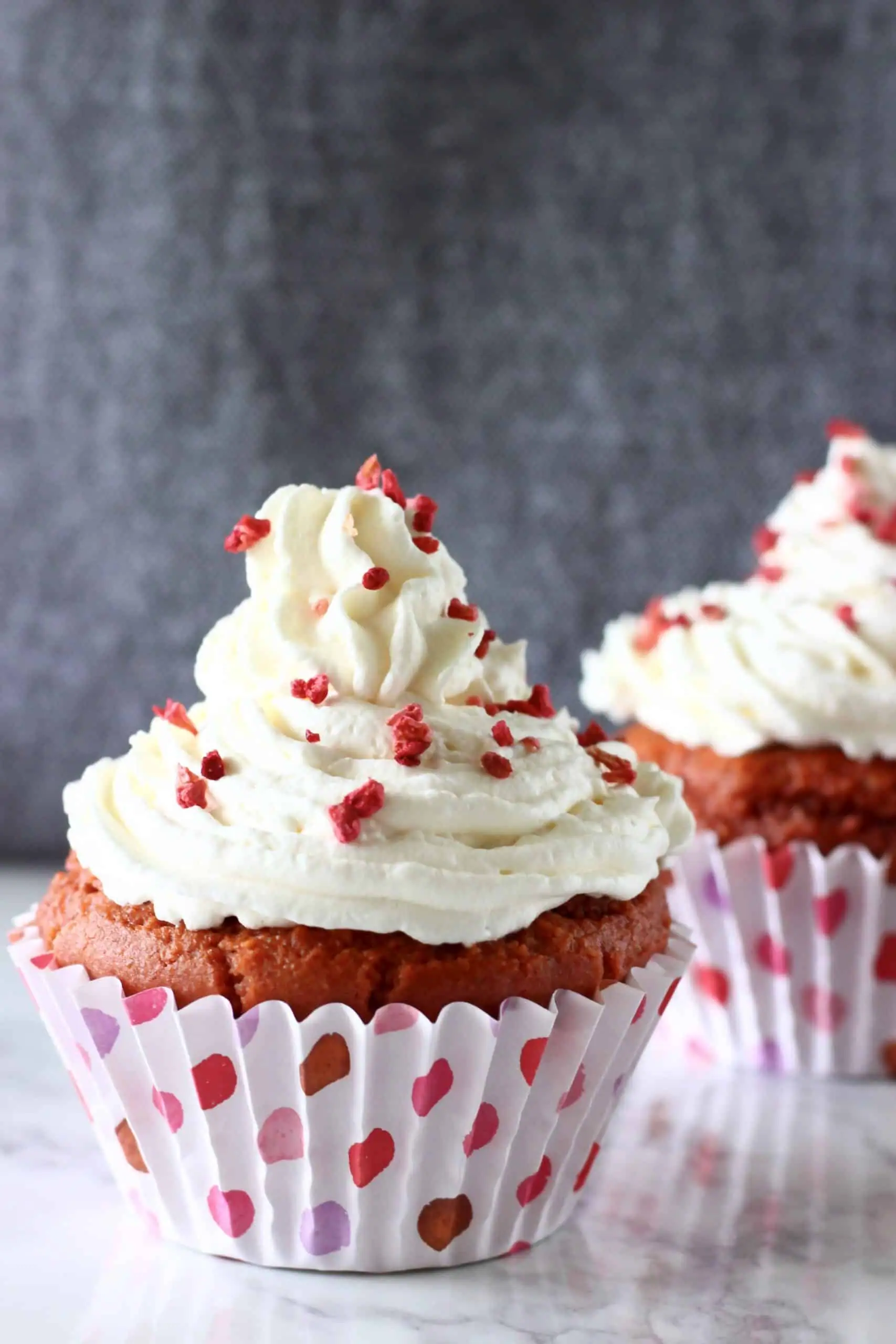 A red velvet cupcake topped with white creamy frosting sprinkled with freeze-dried raspberries on a sheet of brown baking paper