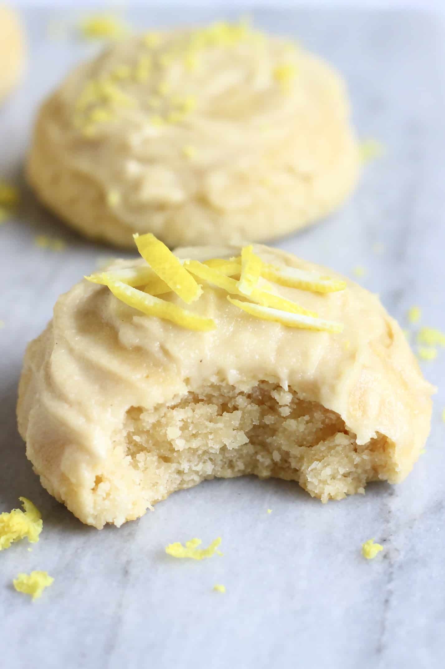 Two gluten-free vegan lemon cookies topped with frosting with a bite taken out of one