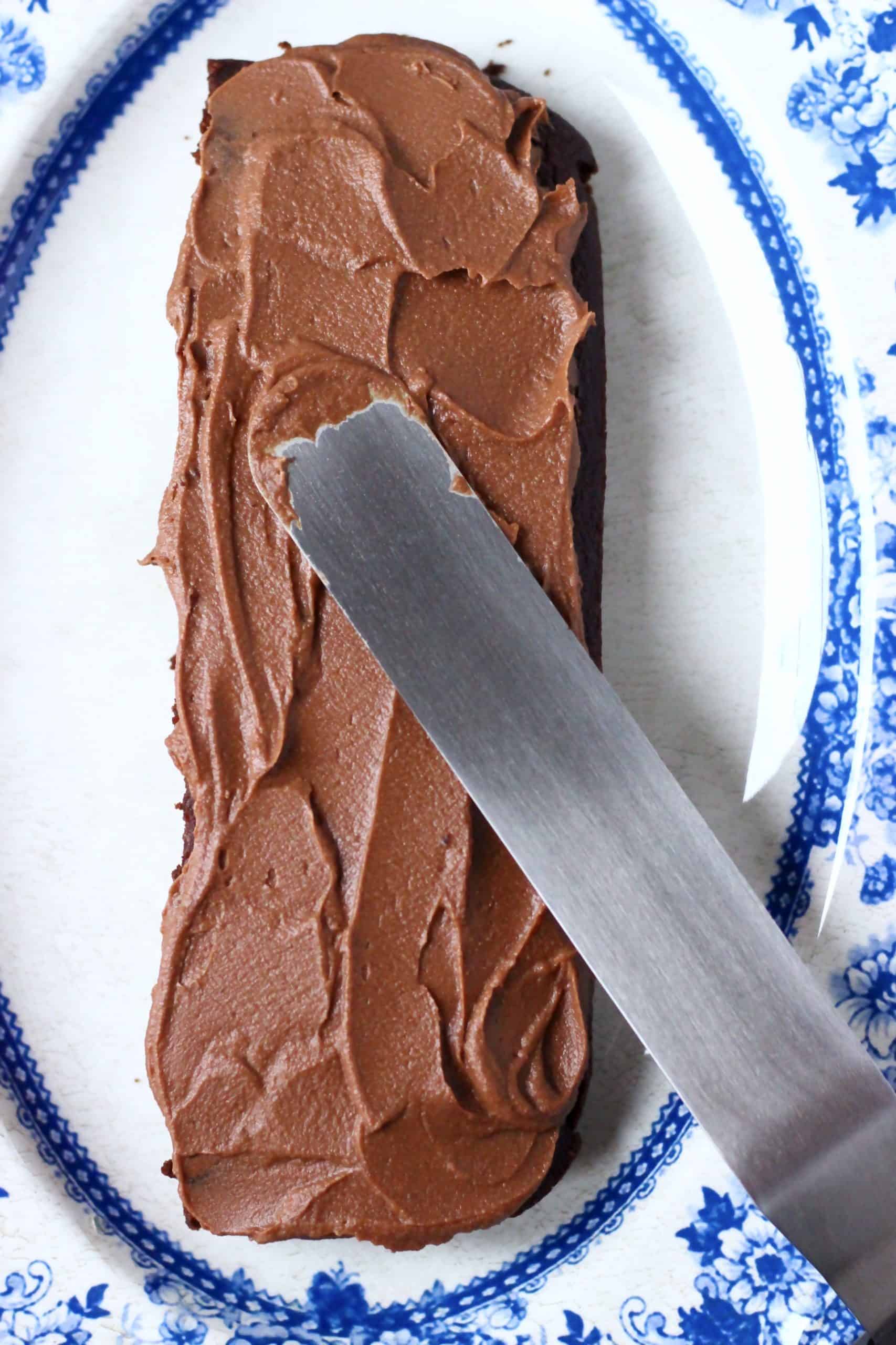 A rectangle of vegan chocolate cake with a palette knife spreading chocolate buttercream on top