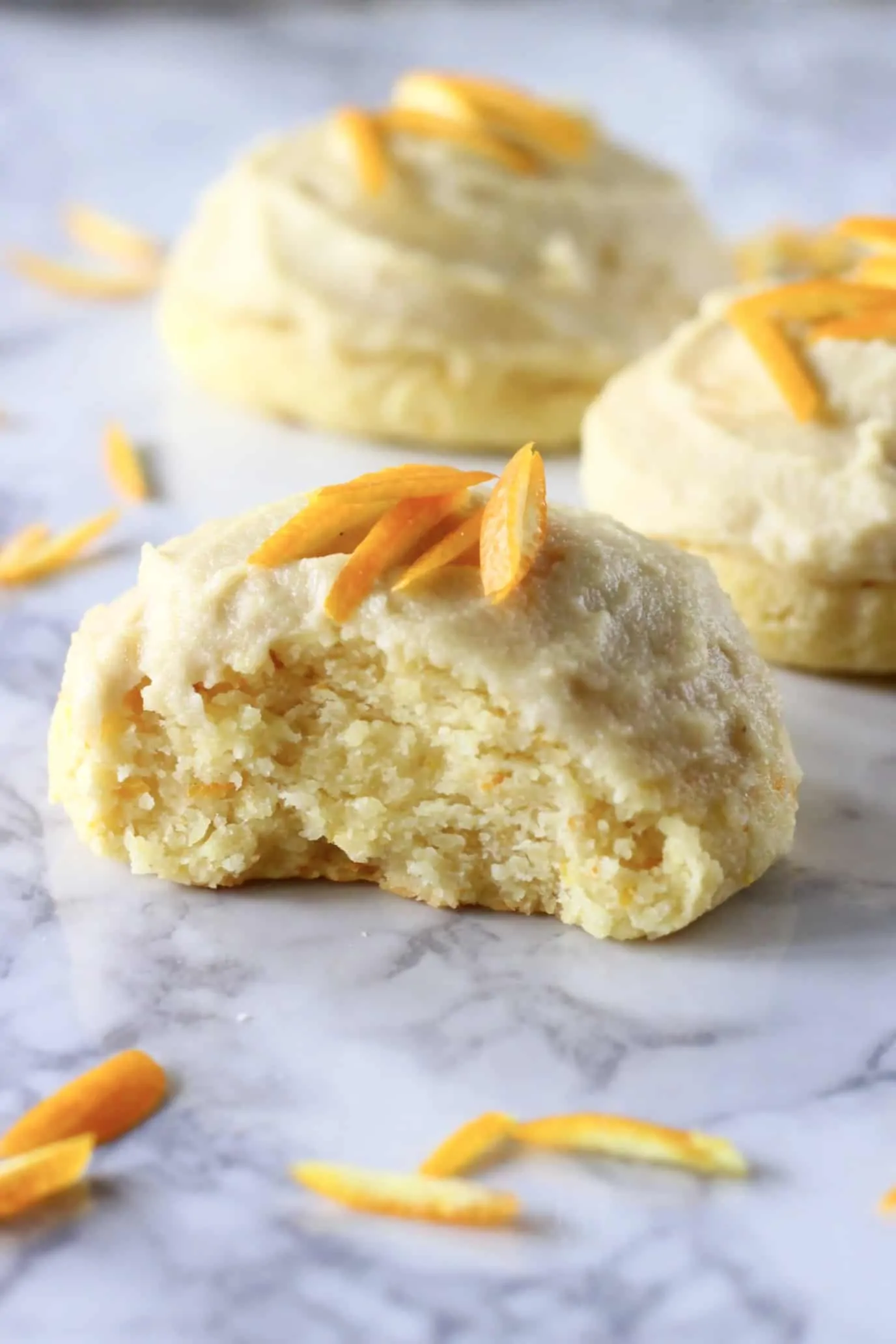 Three gluten-free vegan orange cookies with frosting and orange zest with a bite taken out of one
