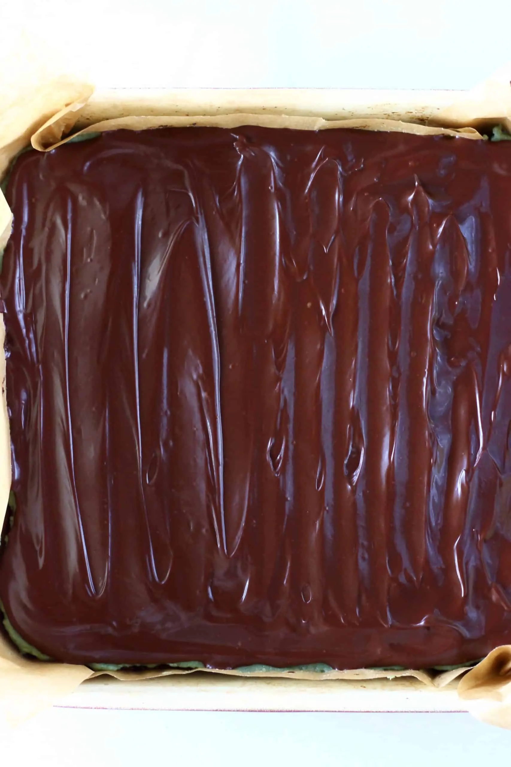 Vegan chocolate ganache spread over vegan peppermint brownies in a square baking tin lined with baking paper