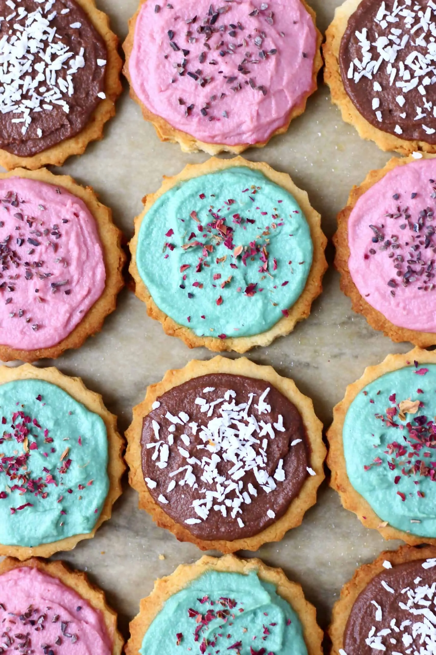 Twelve circular sugar cookies topped with different coloured frosting and sprinkles on a sheet of brown baking paper