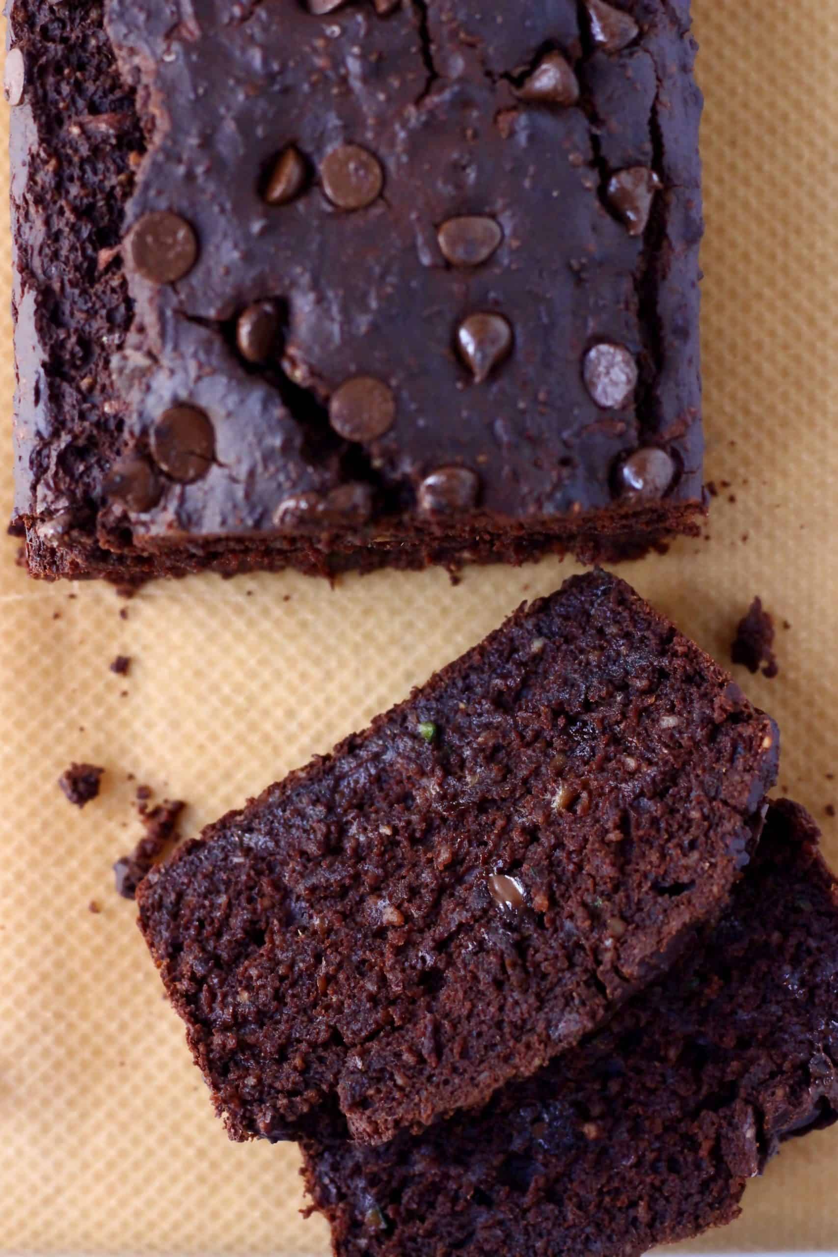 A loaf of chocolate zucchini bread studded with chocolate chips with two slices taken from it against a sheet of brown baking paper