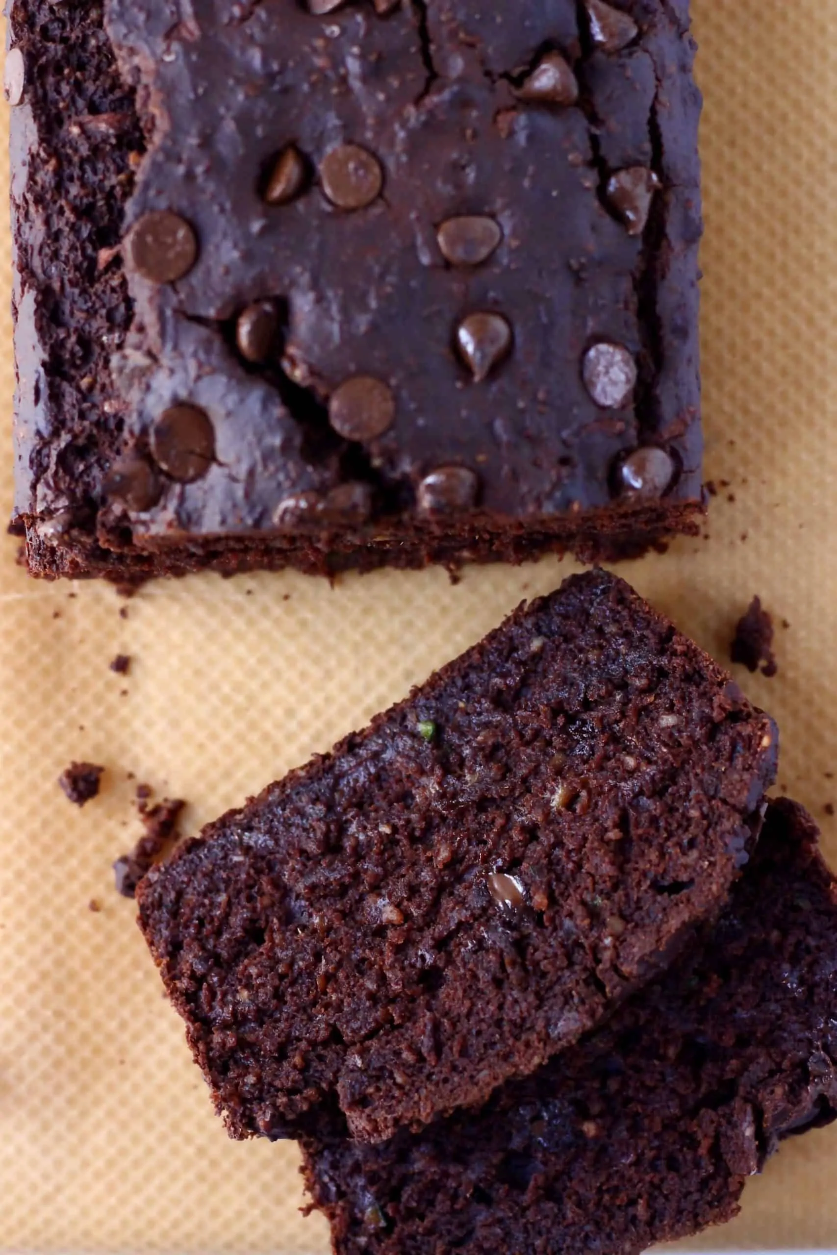 A loaf of chocolate zucchini bread studded with chocolate chips with two slices taken from it against a sheet of brown baking paper