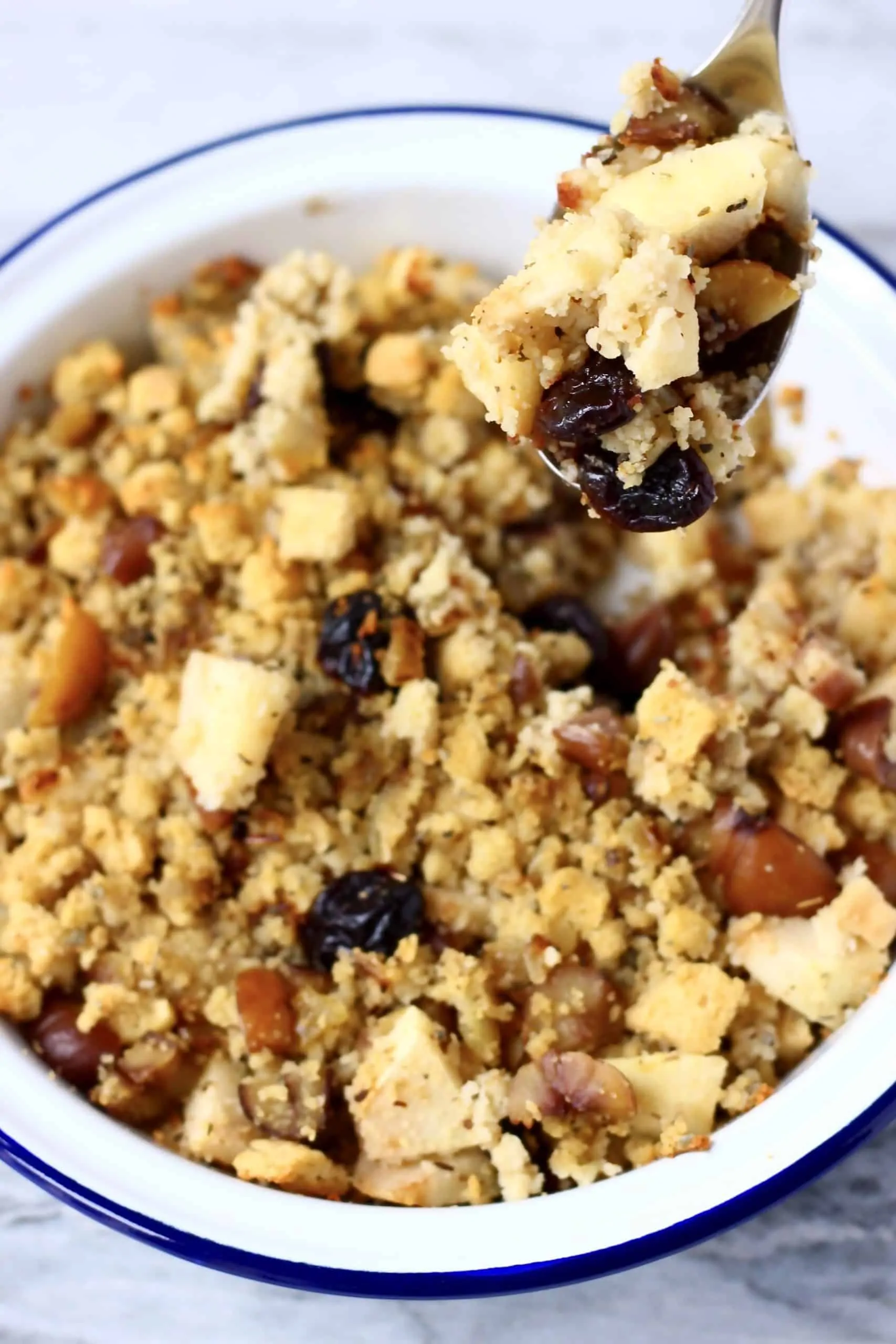 Stuffing with chestnuts, apple pieces and dried cherries in a white pie dish with a blue rim with a silver spoon holding up a mouthful of stuffing