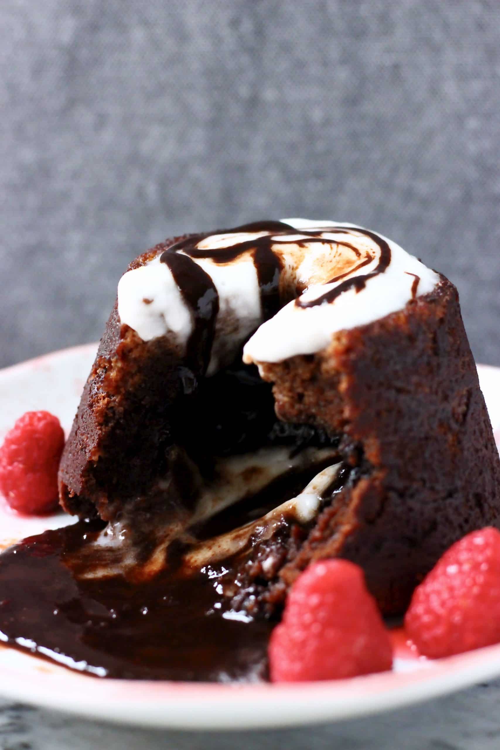 Chocolate lava cake on a plate with chocolate sauce flowing out of it topped with white cream and decorated with raspberries against a grey background