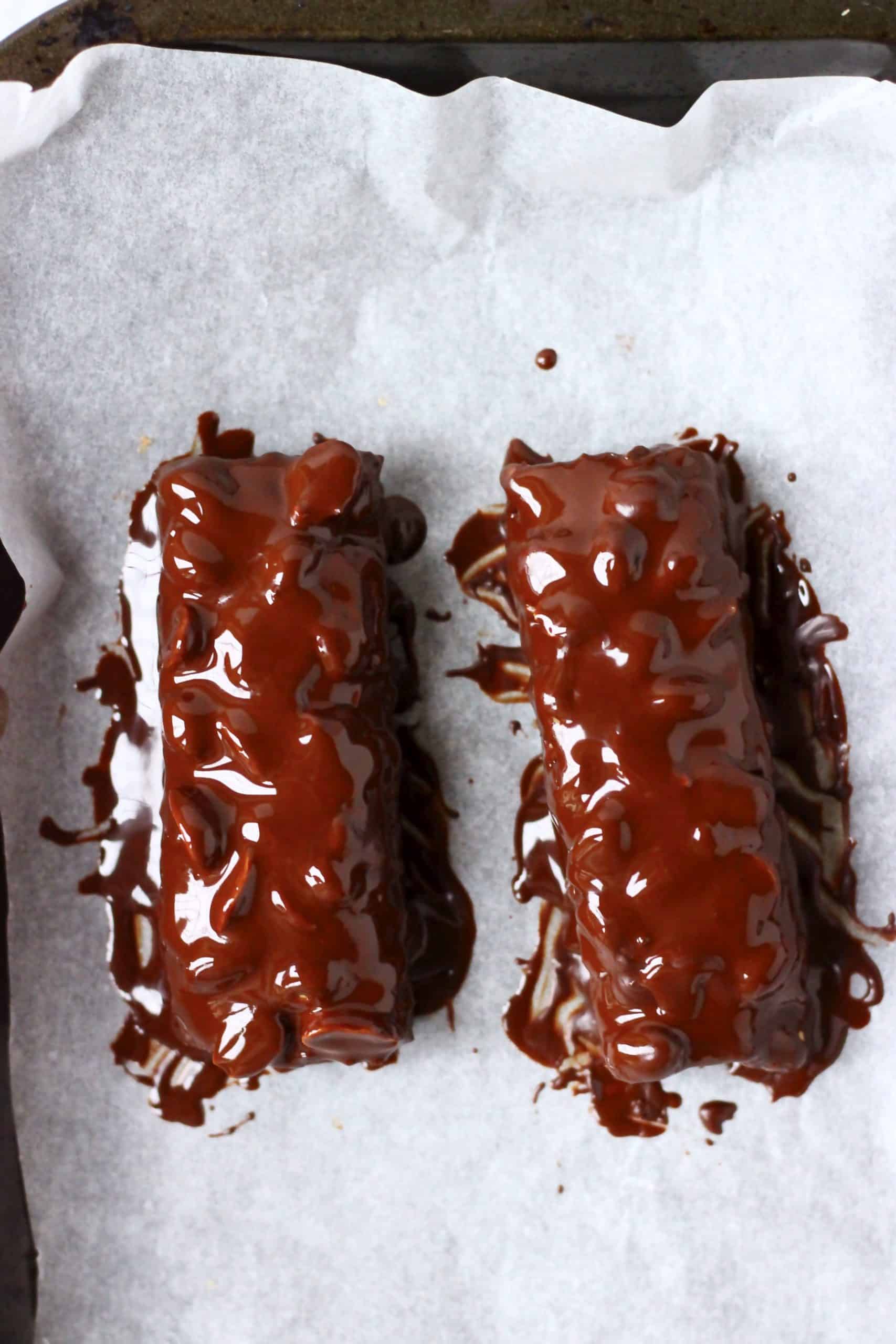 Two vegan snickers bars dipped in melted chocolate on a baking tray lined with paper