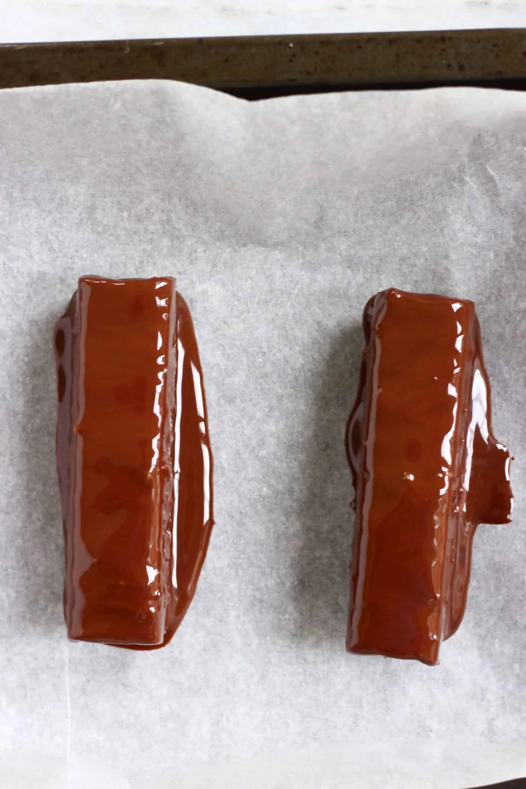 Two vegan twix bars dipped in melted chocolate on a tray lined with baking paper