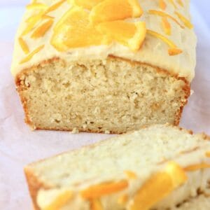 A loaf of vegan orange bread topped with frosting and orange slices with two slices next to it