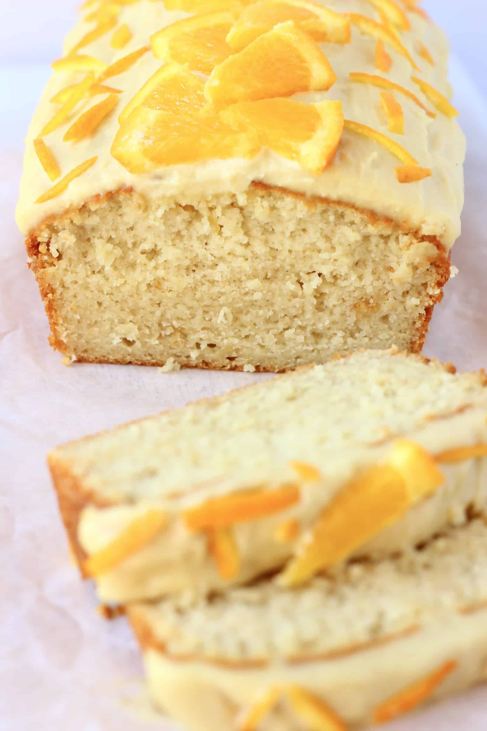A loaf of vegan orange bread topped with frosting and orange slices with two slices next to it