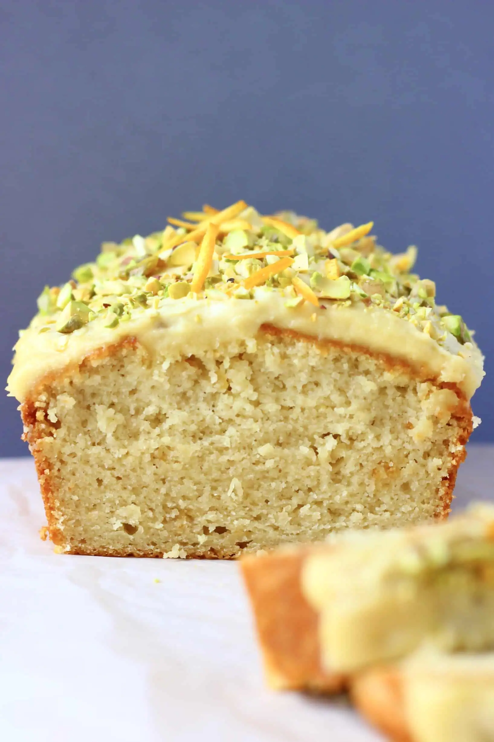A loaf of vegan orange bread topped with frosting and chopped pistachios with two slices next to it