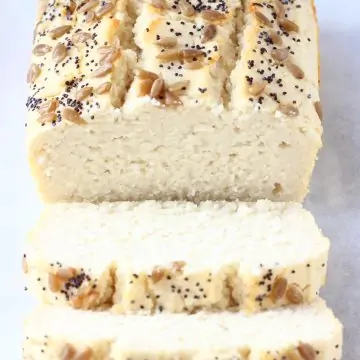 A loaf of coconut flour bread topped with sunflower seeds and poppy seeds with two slices next to it