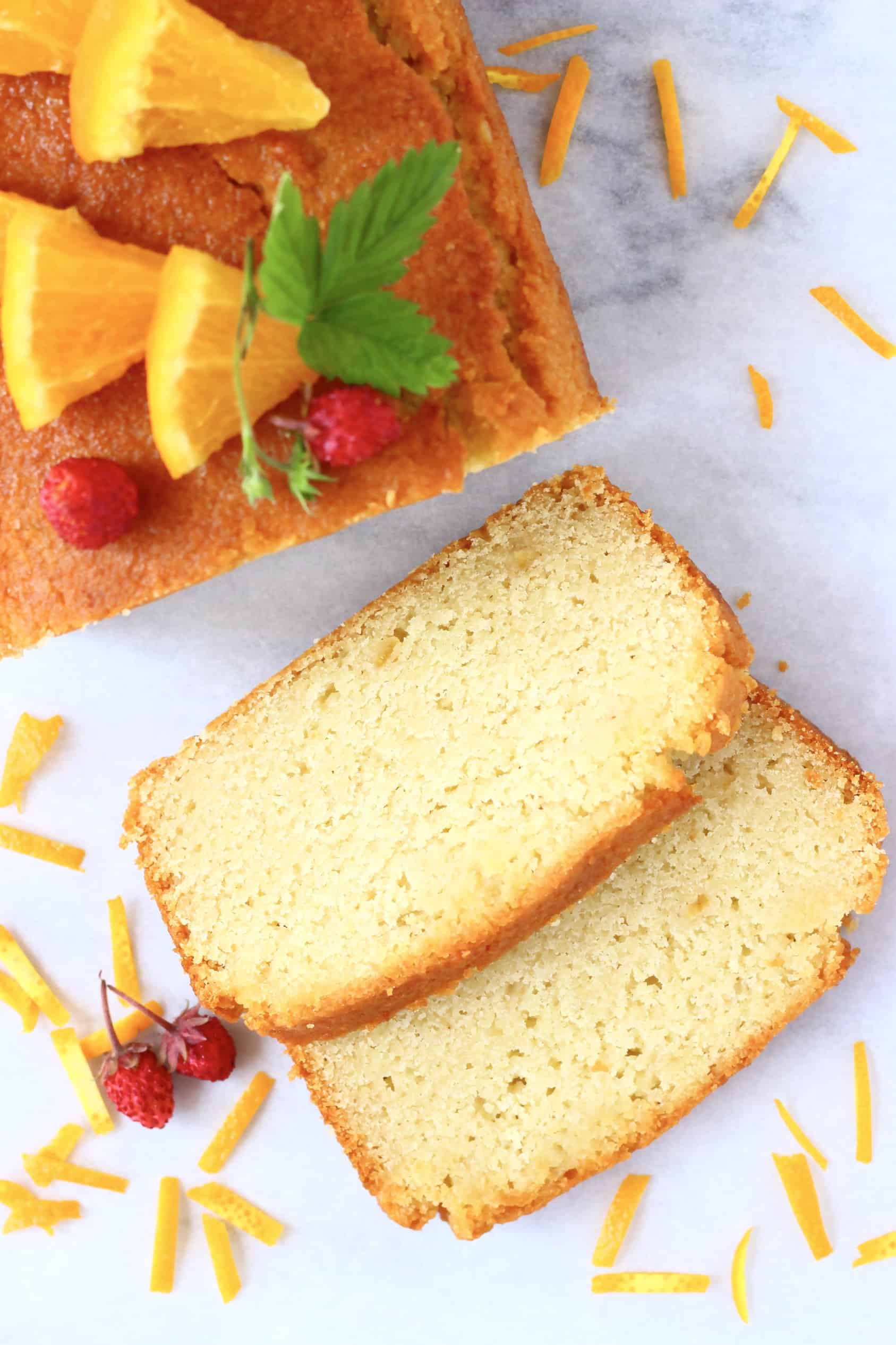 A loaf of gluten-free vegan orange drizzle cake topped with mini strawberries and orange slices with two slices next to it
