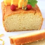 A loaf of gluten-free vegan orange drizzle cake topped with mini strawberries and orange slices with two slices next to it