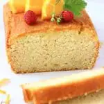 A loaf of gluten-free vegan orange drizzle cake topped with mini strawberries and orange slices with two slices next to it