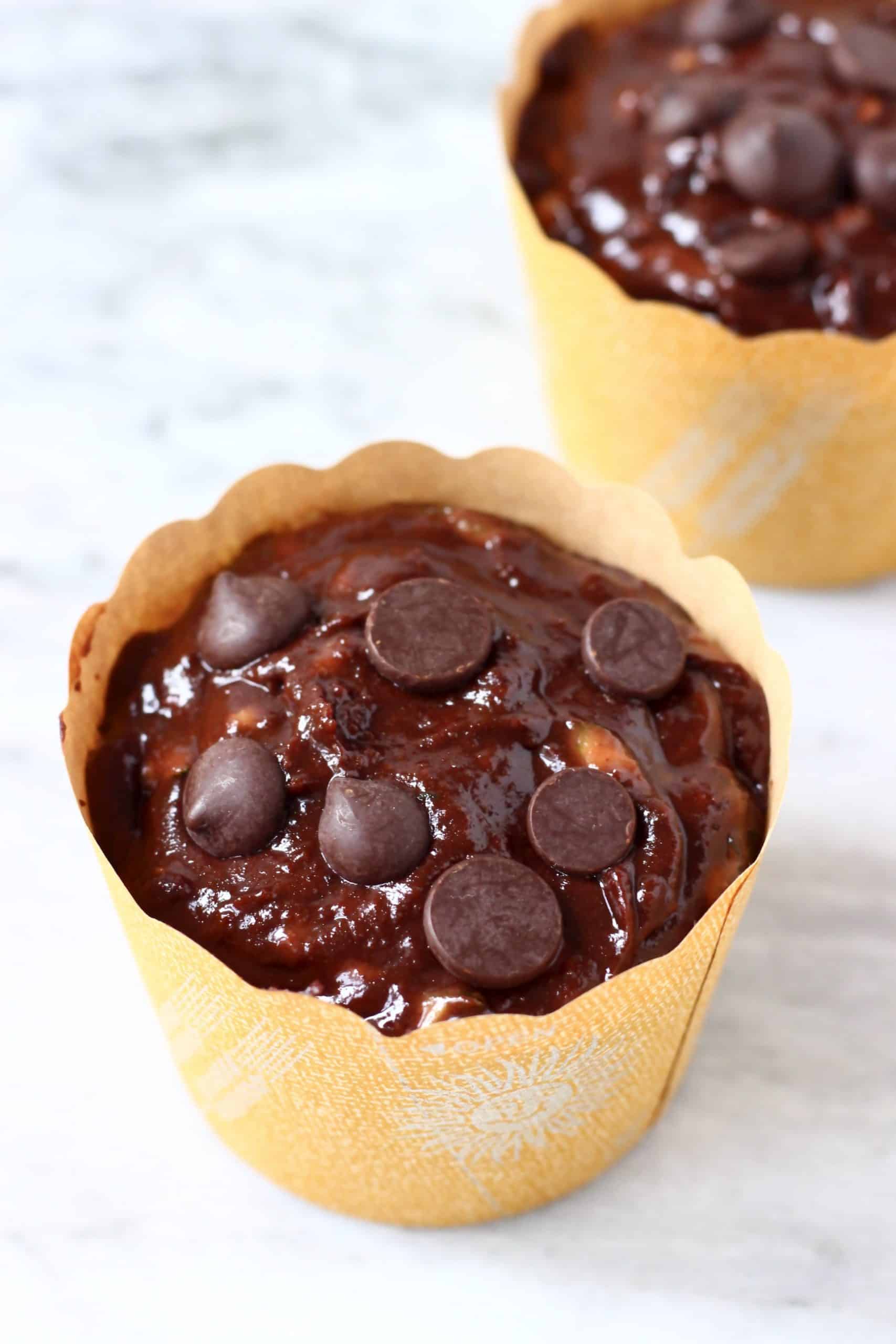 Two brown muffin cases filled with chocolate zucchini muffin batter and chocolate chips
