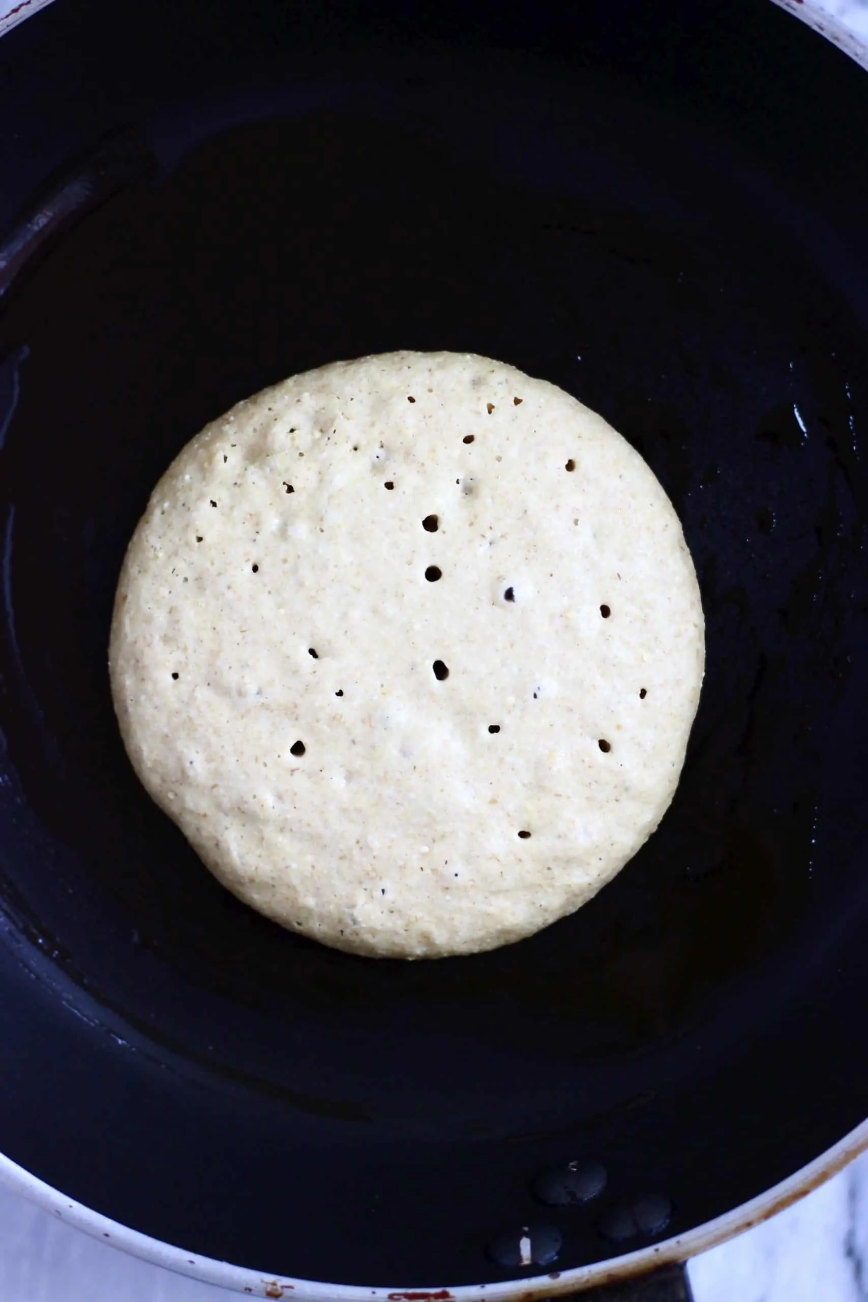 A quinoa pancake with bubbles on the surface being cooked in a black frying pan