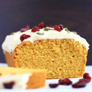 A sliced gluten-free vegan pumpkin loaf cake topped with cream cheese frosting, dried cranberries and pumpkin seeds