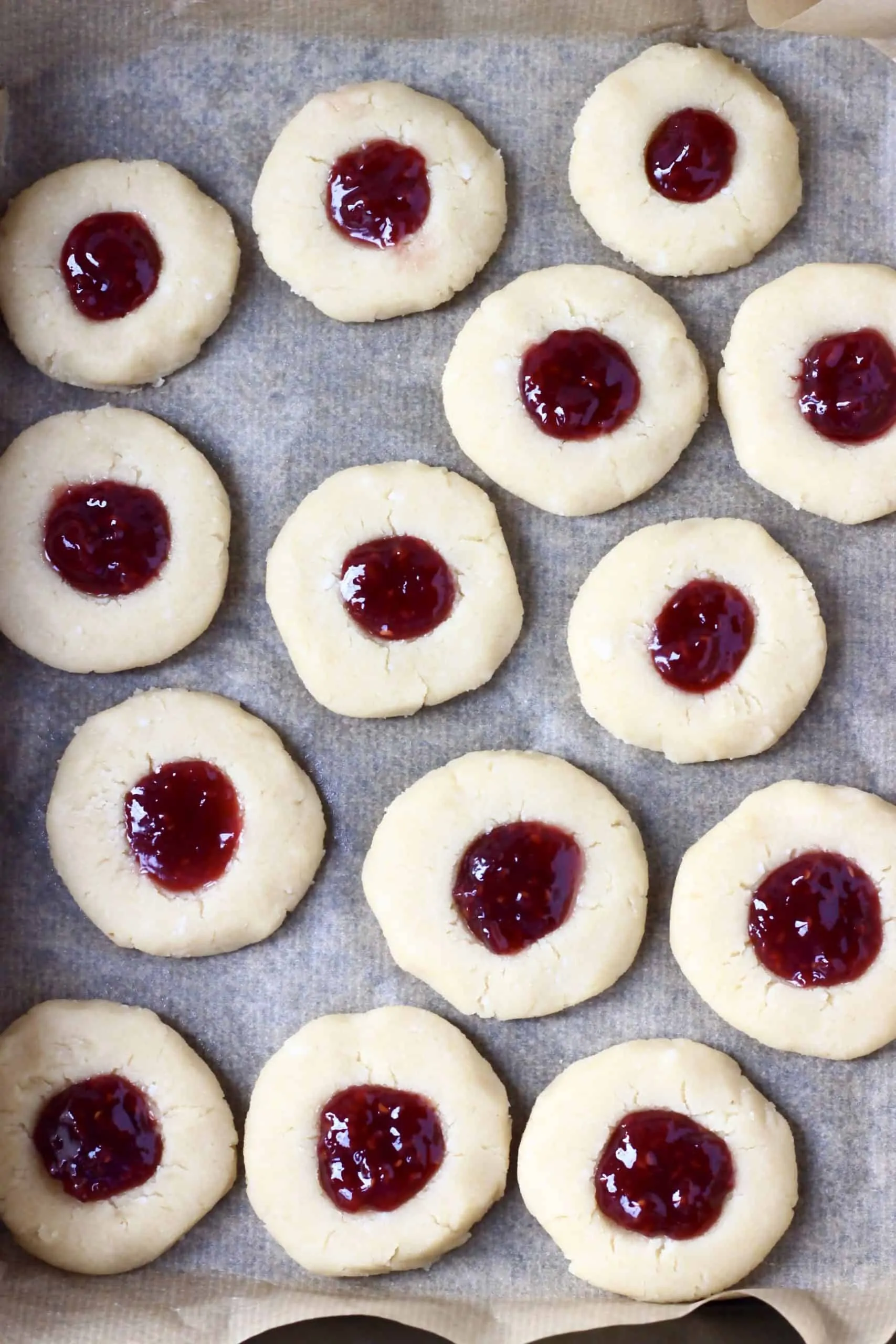 Raw gluten-free vegan thumbprint cookies with indents filled with raspberry jam on a baking tray lined with baking paper