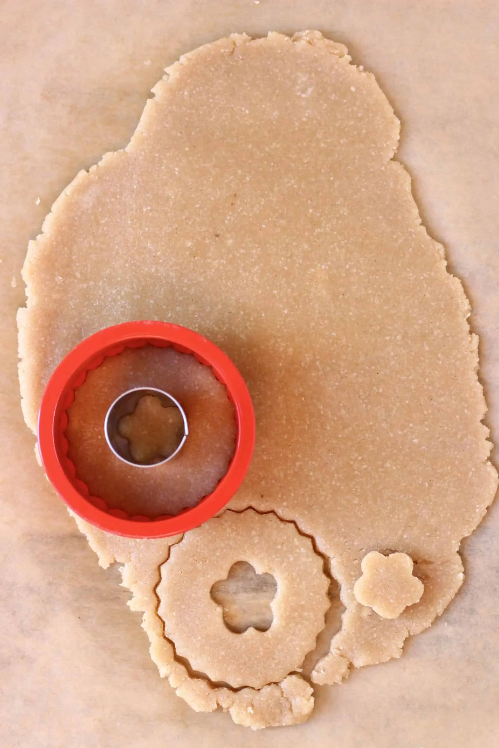 Raw gluten-free vegan linzer cookie dough rolled out on a sheet of baking paper with a circular cookie cutter and a flower-shaped cookie cutter cutting out shapes