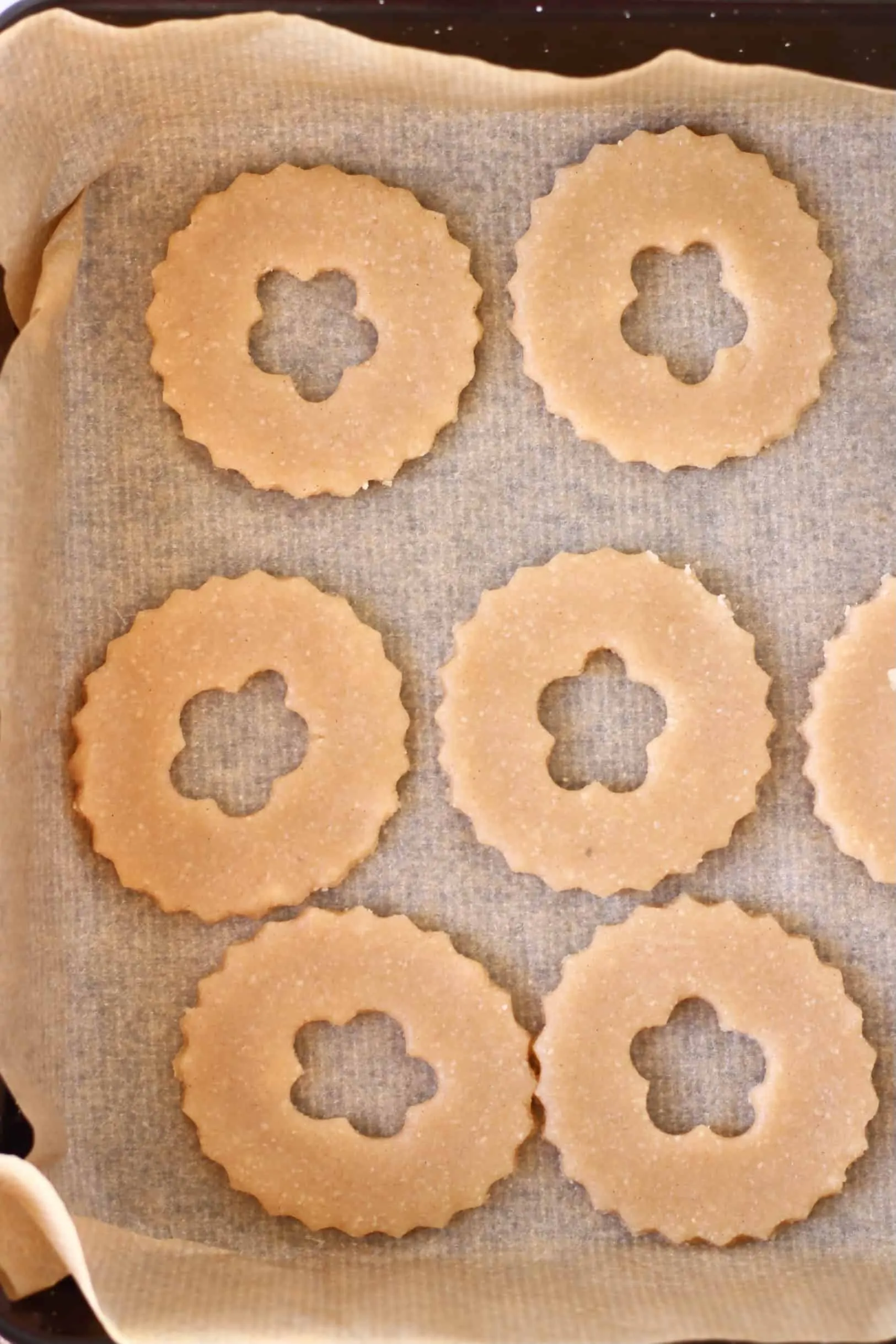 Six raw gluten-free vegan linzer cookie circles with flower-shaped windows on a baking tray lined with baking paper