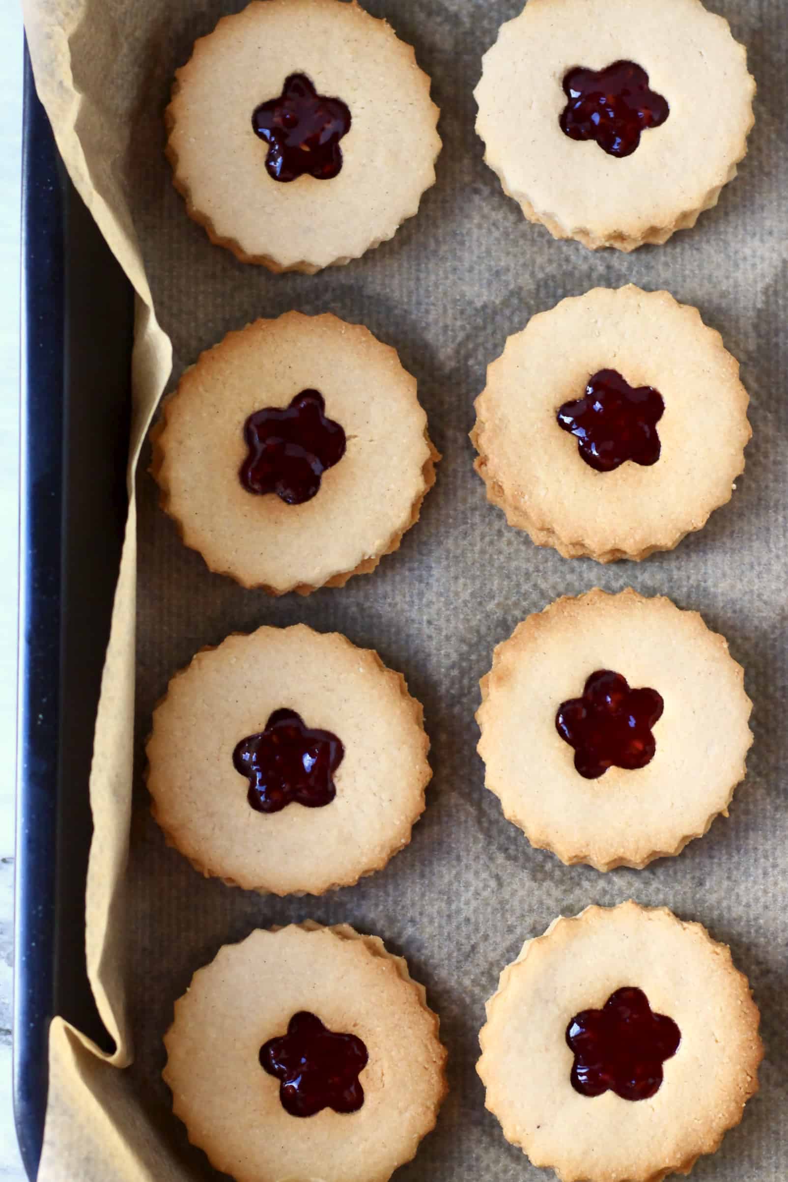 Eight gluten-free vegan linzer cookies sandwiched with raspberry jam on a baking tray lined with baking paper