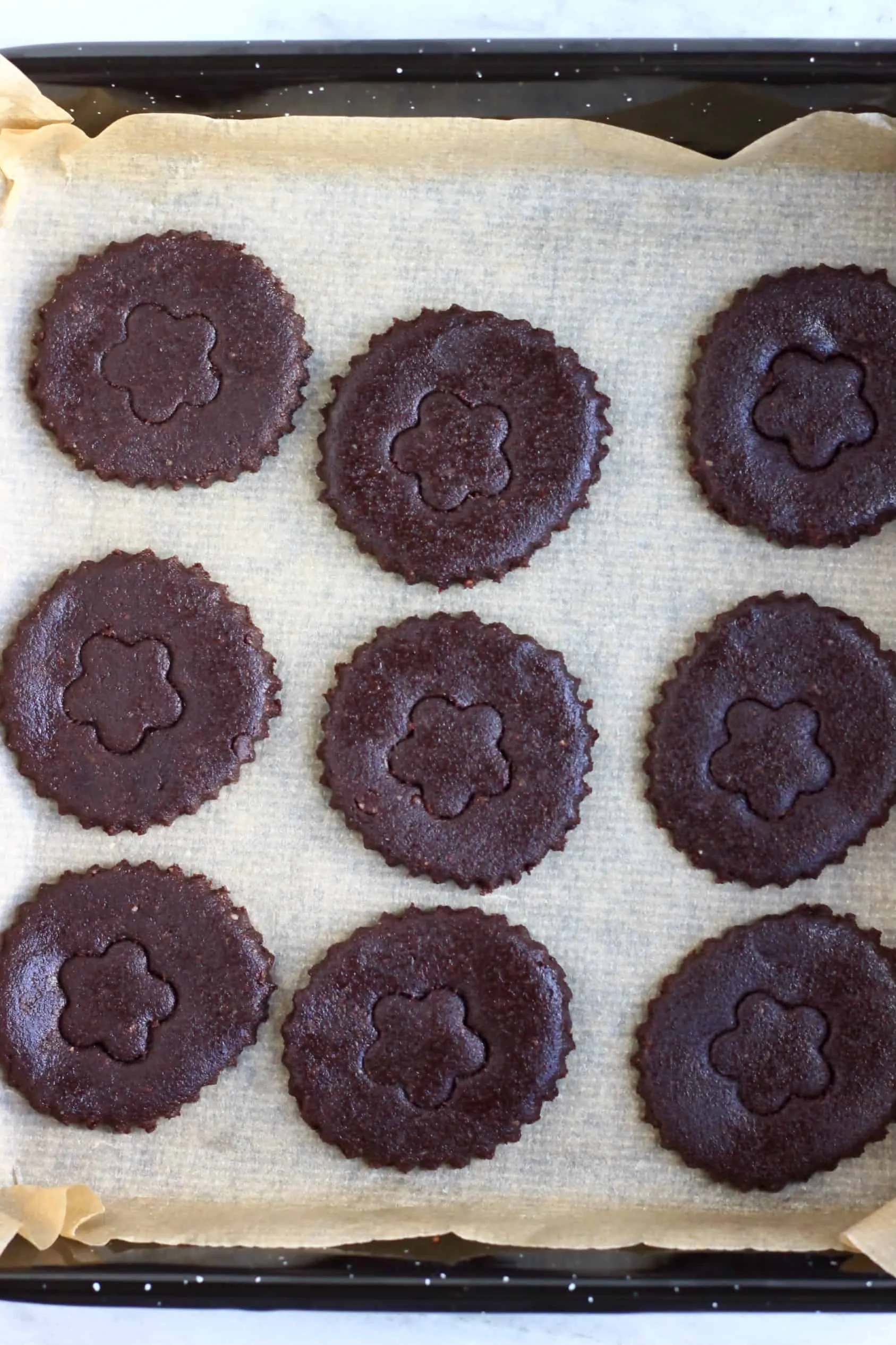 Nine raw gluten-free vegan homemade oreo cookie circles decorated with flower stamps on a baking tray lined with baking paper