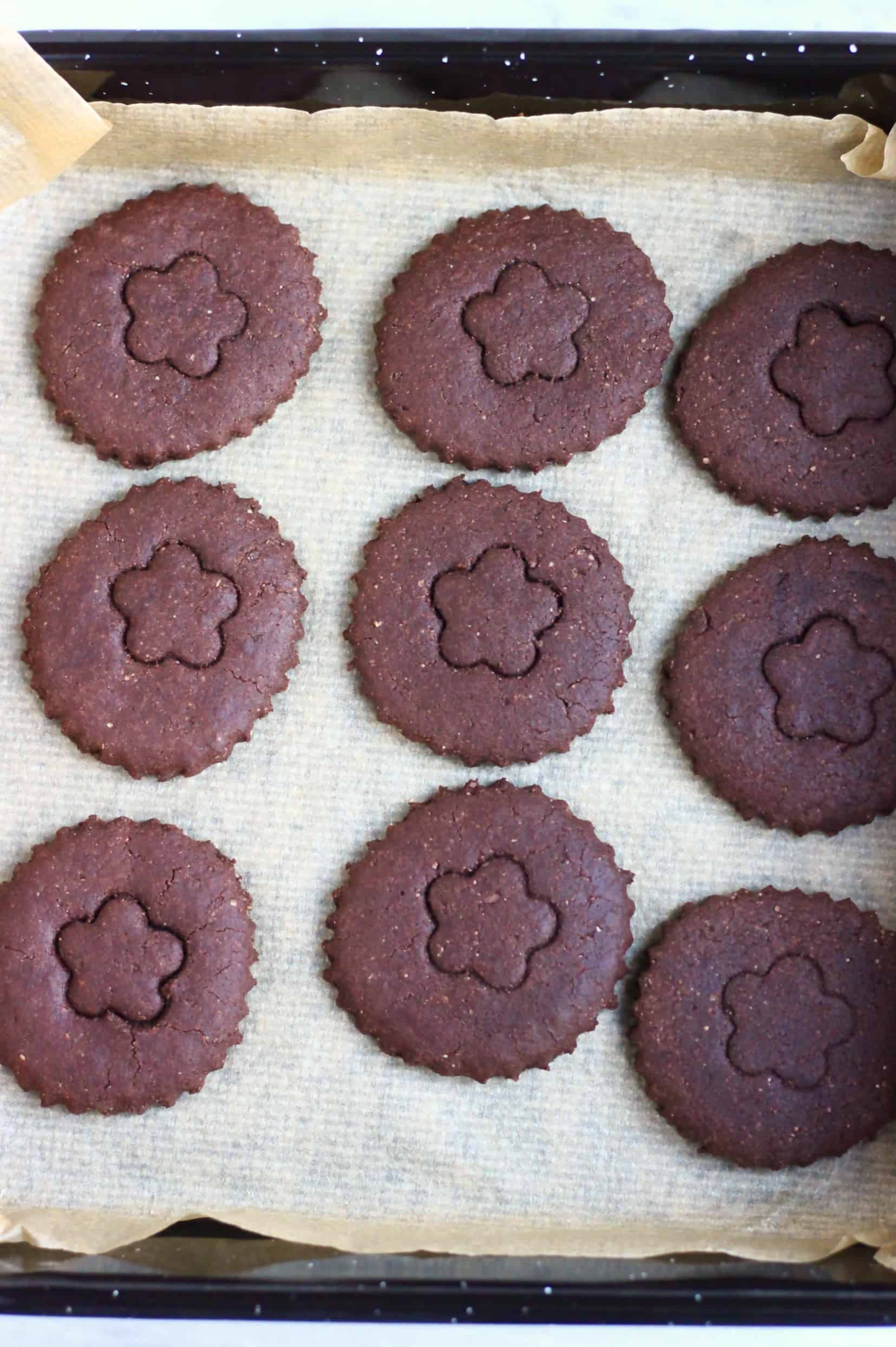 Nine gluten-free vegan homemade oreo cookie circles decorated with a flower pattern on a baking tray lined with baking paper