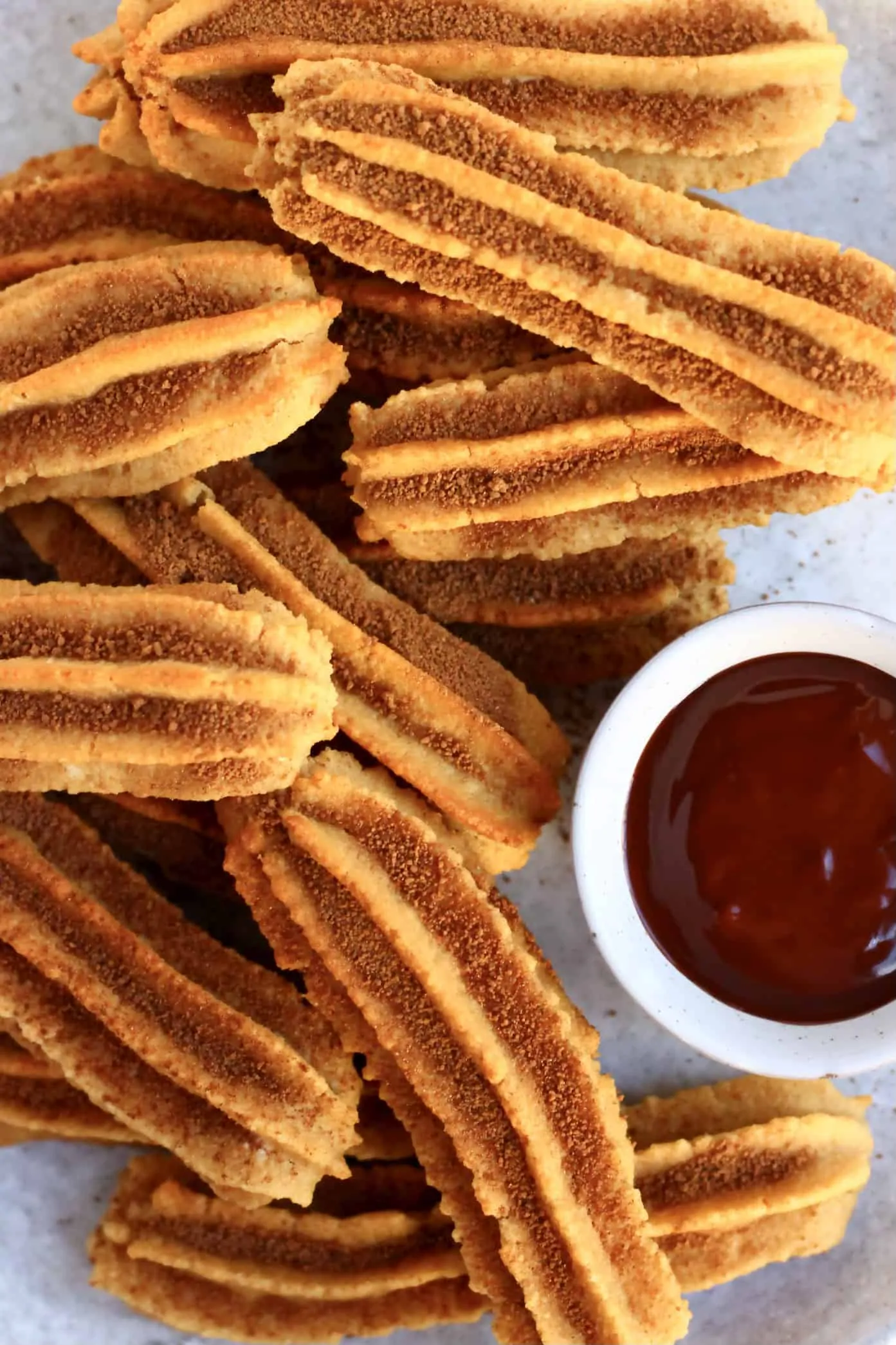 Several gluten-free vegan churros dusted with cinnamon sugar on a plate with a bowl of vegan chocolate sauce