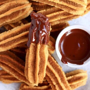 Several gluten-free vegan churros on a plate with a pot of vegan chocolate sauce and one of them dipped in it