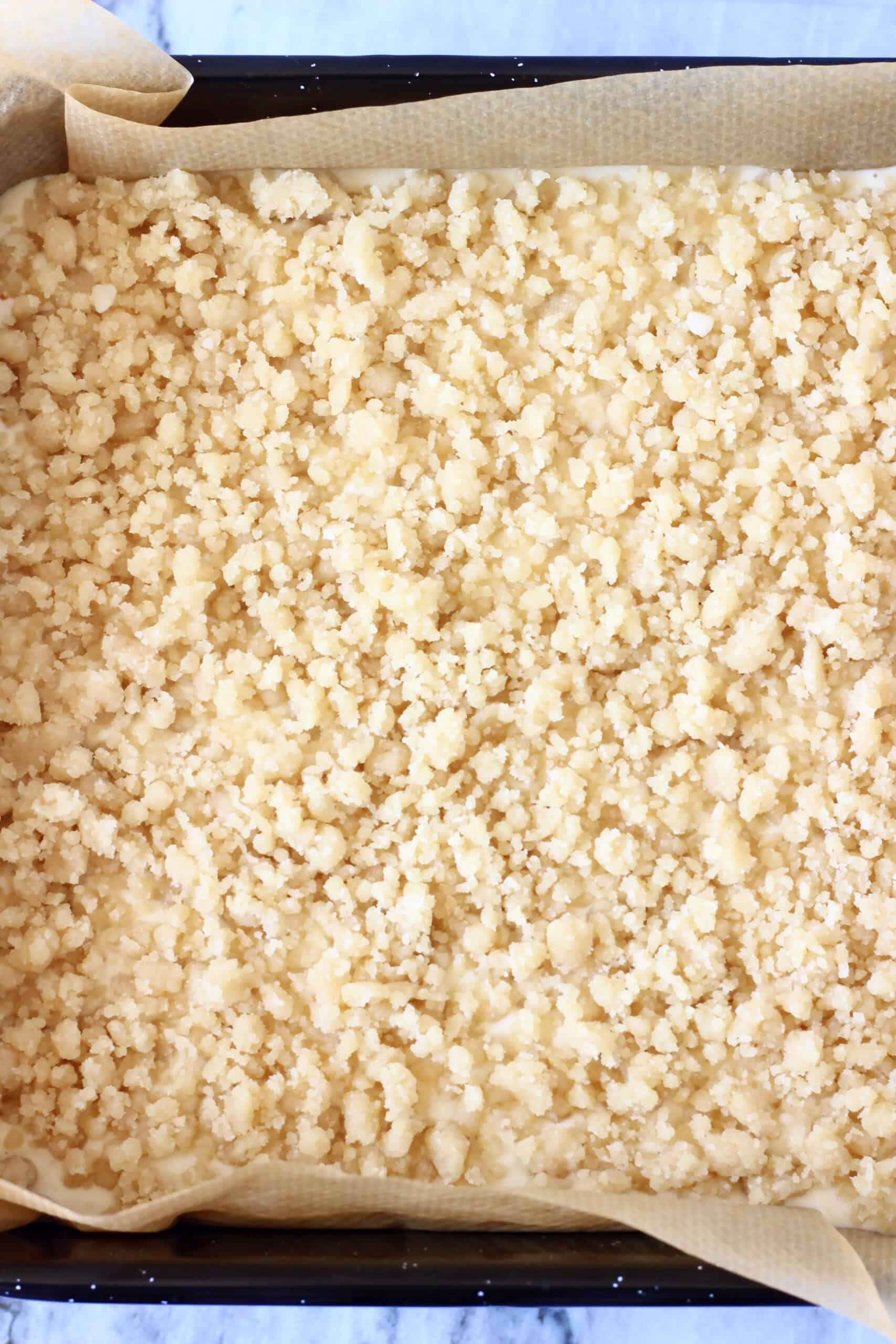Raw crumble topping dough sprinkled over vegan cheesecake filling in a square baking tin lined with baking paper