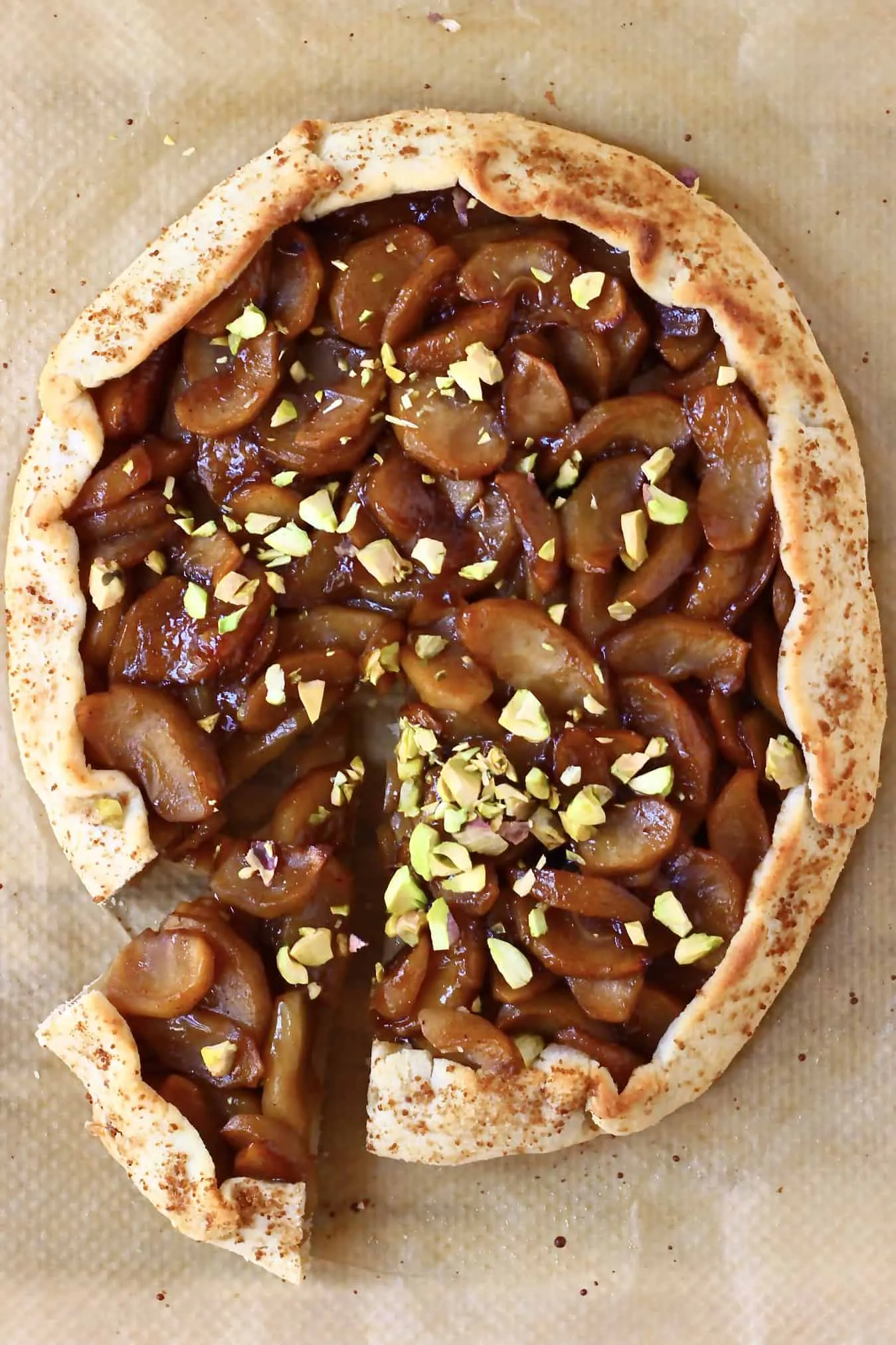 A round galette filled with brown apples sprinkled with pistachio nuts with a slice cut out of it against a sheet of brown baking paper