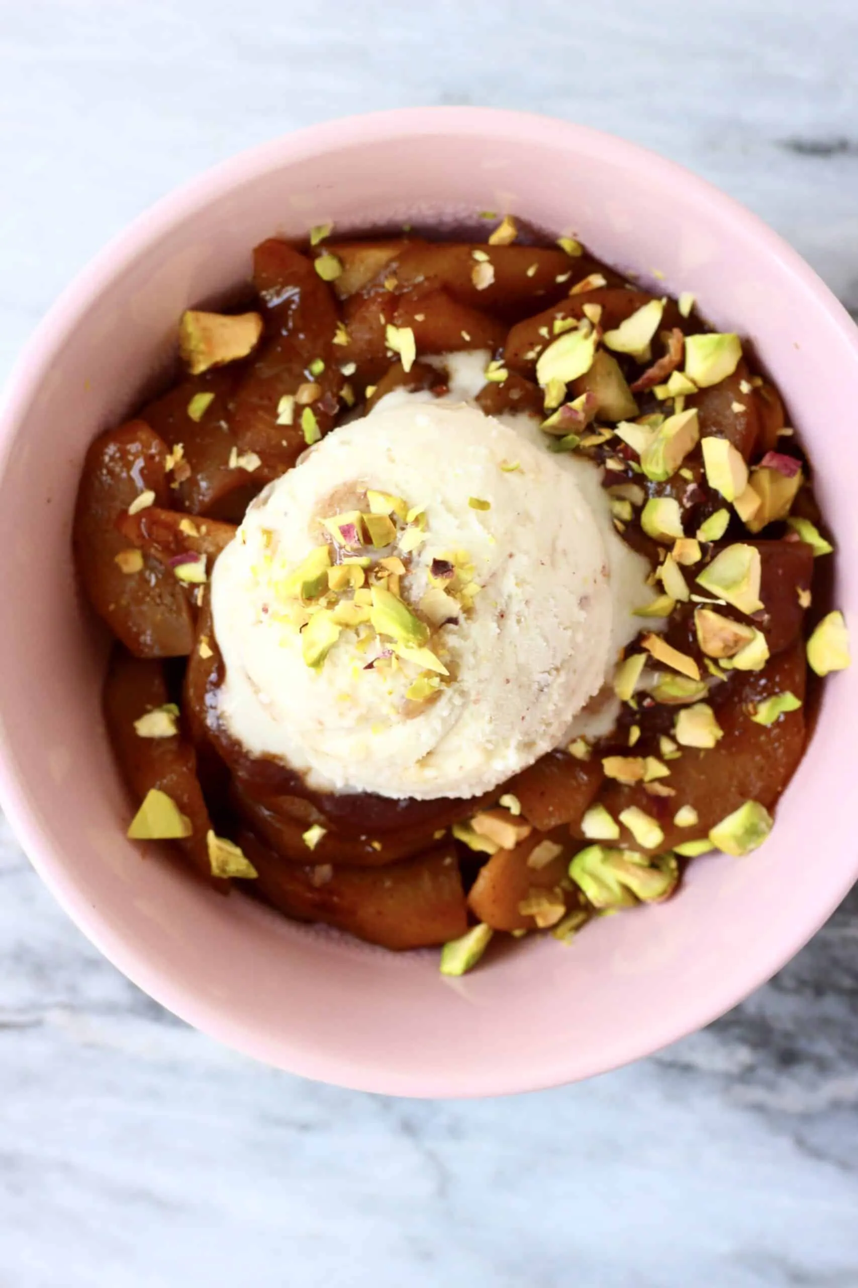 Sliced baked apples in a bowl scattered with chopped pistachio nuts and topped with ice cream