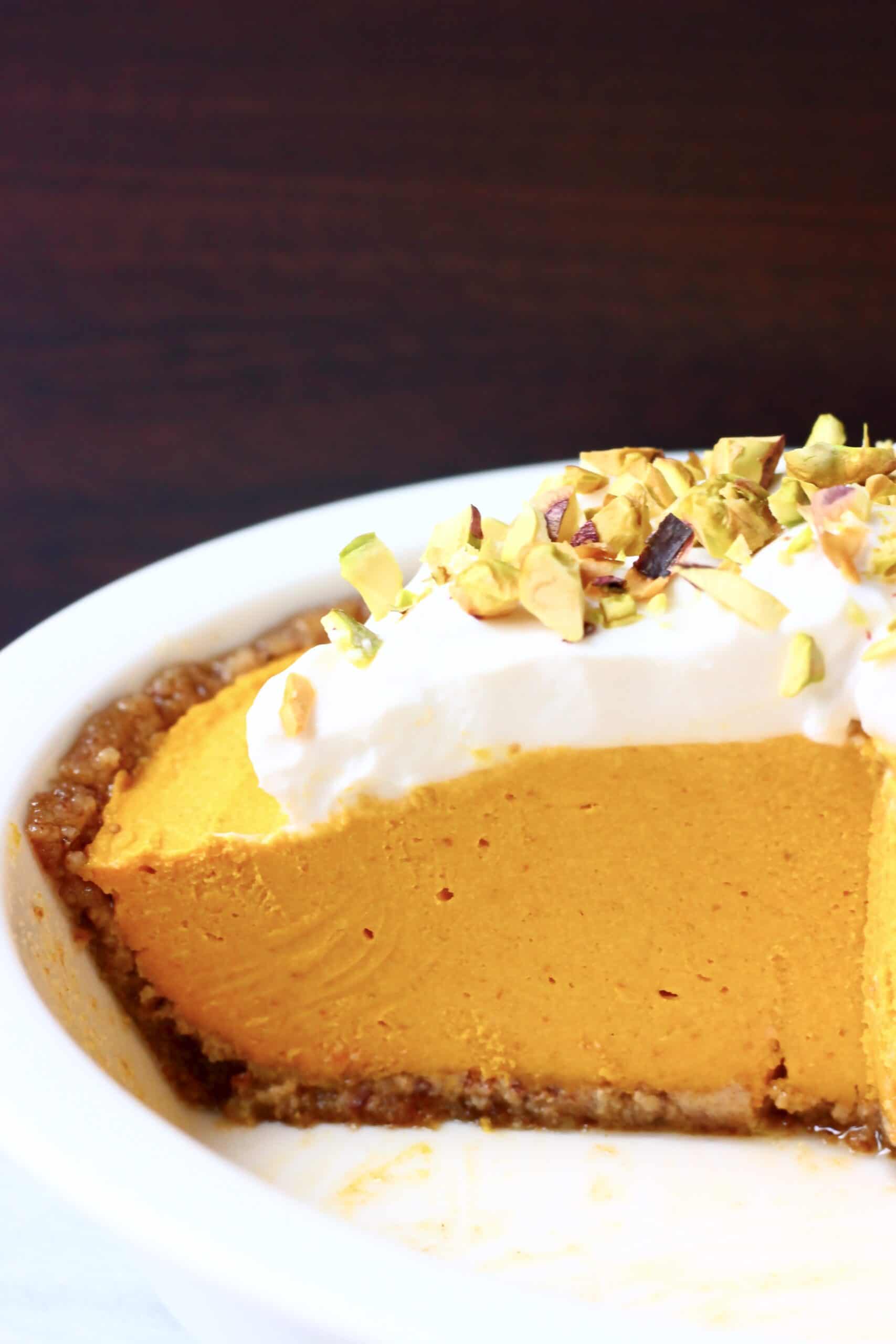 A white pie dish with vegan no-bake pumpkin pie topped with whipped cream and chopped pistachios against a dark brown background