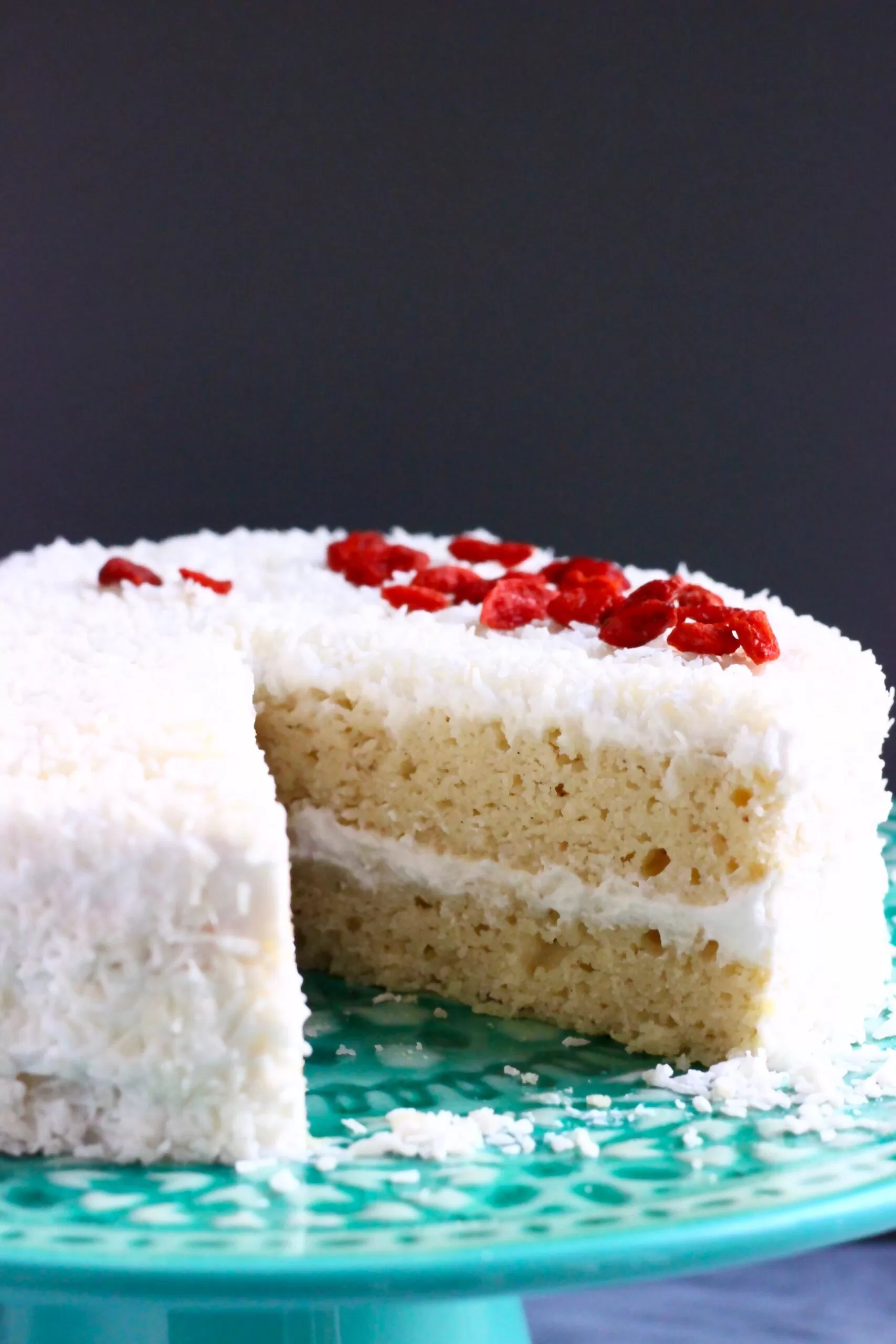A white sponge cake covered in creamy frosting and coconut sprinkled with red goji berries against a dark grey background