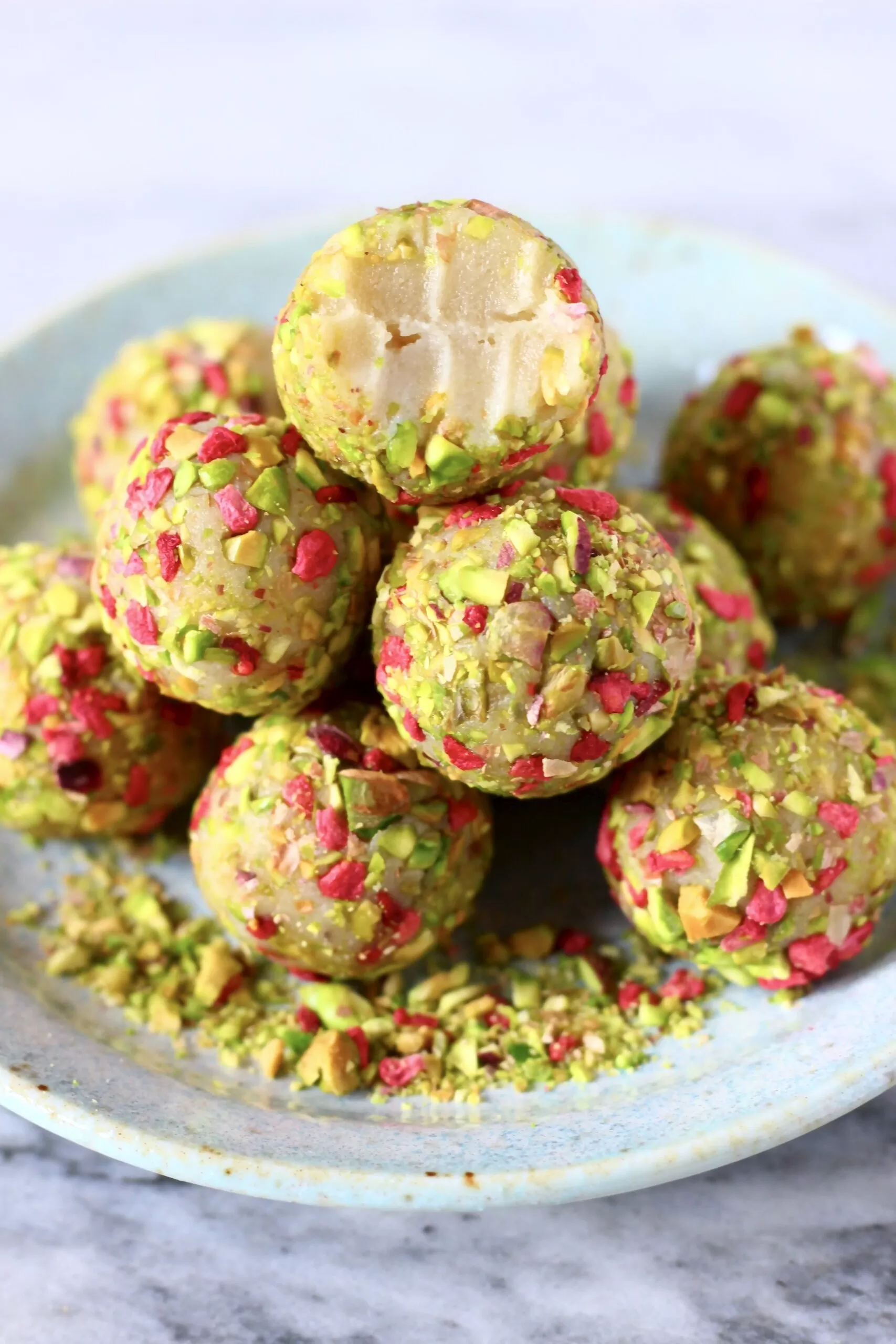 A pile of white chocolate truffles covered in chopped pistachios and freeze-dried raspberries with a bitten one on top on a light blue plate against a marble background
