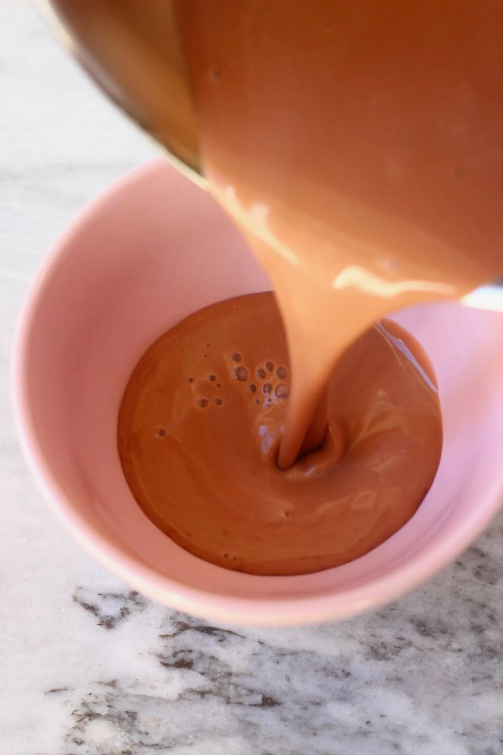 A saucepan pouring hot chocolate into a small pink bowl against a marble background