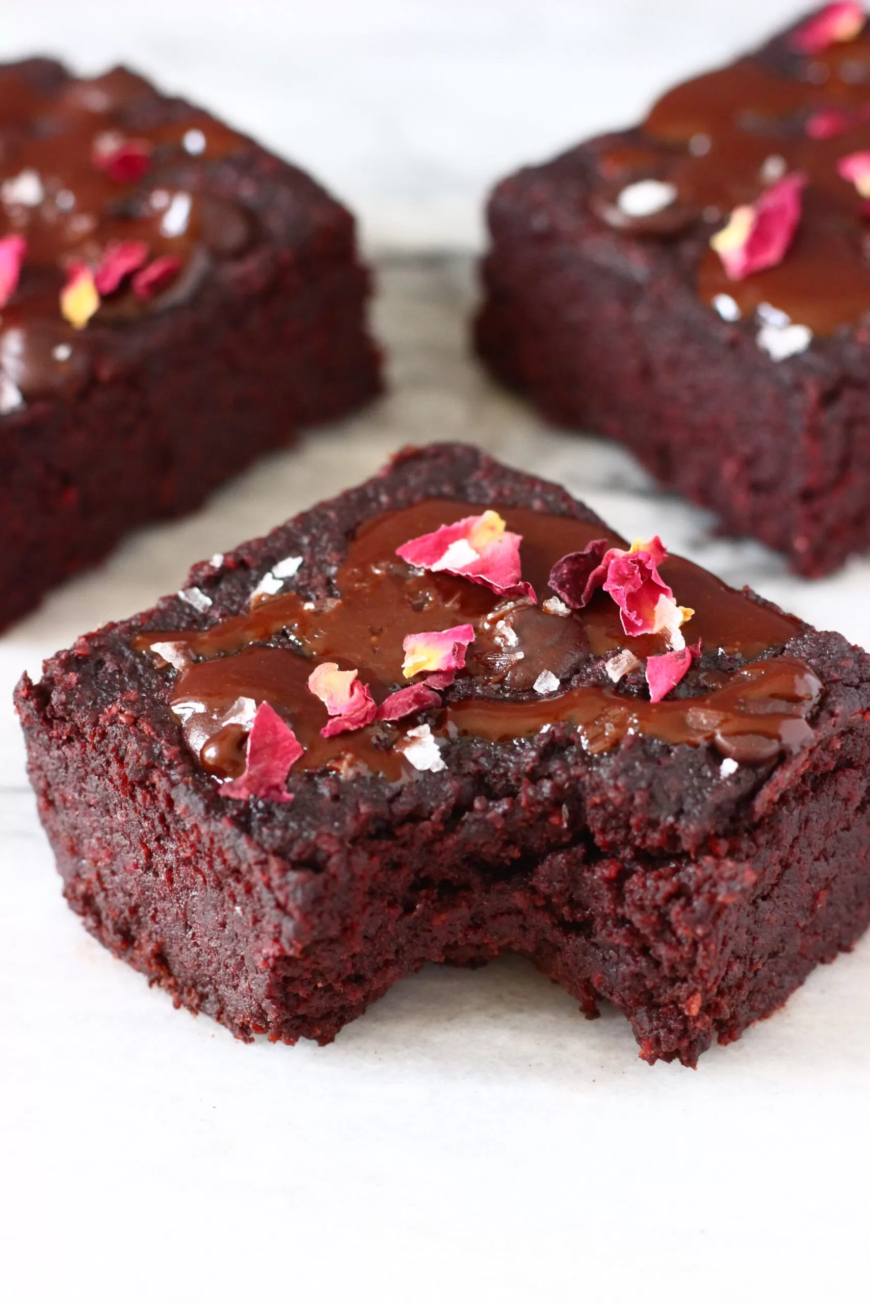 Three beetroot brownies with chocolate frosting, rose petals and salt flakes with a bite taken out of one