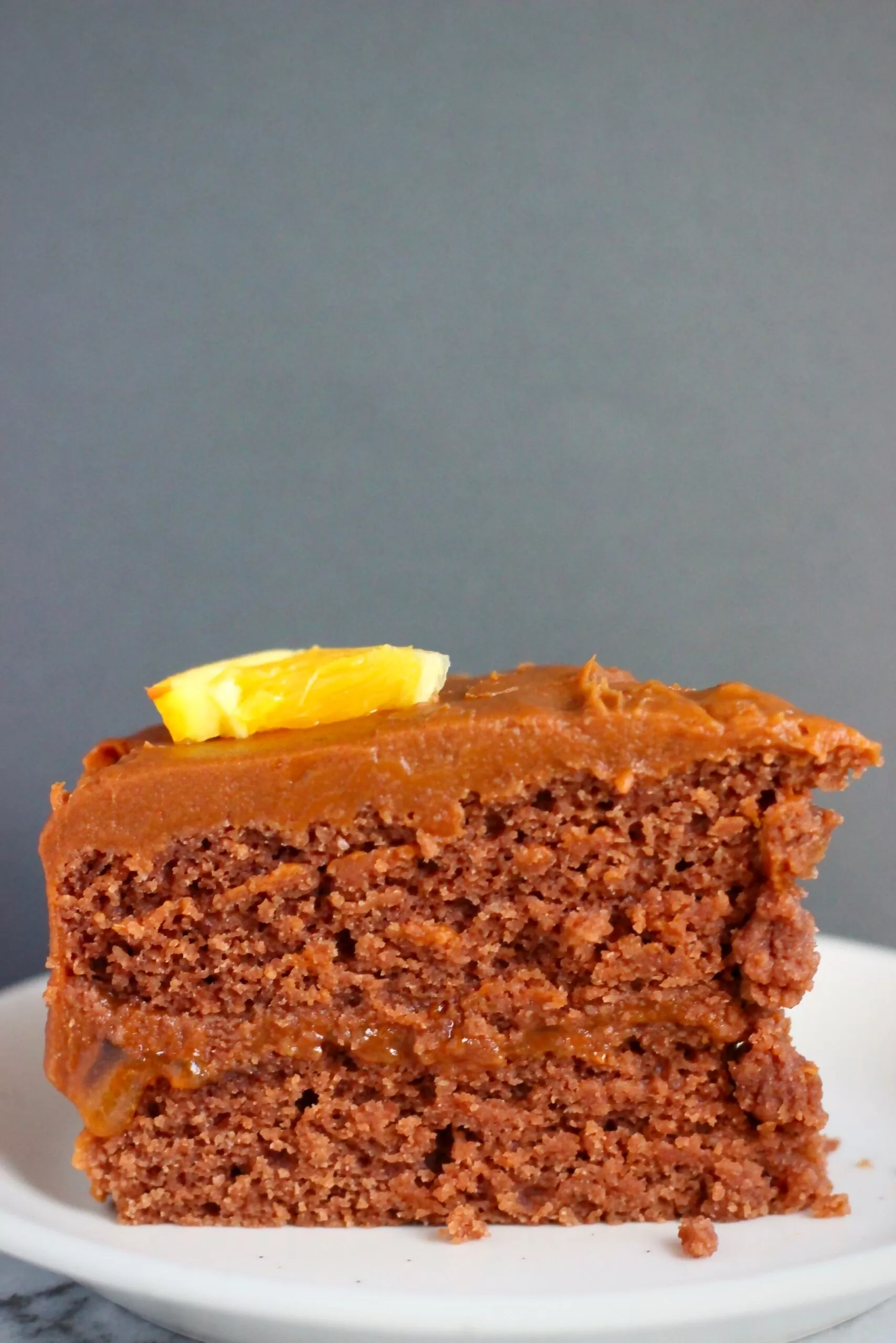 A slice of chocolate sponge sandwiched with chocolate frosting topped with a slice of orange against a dark grey background