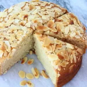 A round gluten-free vegan almond cake topped with flaked almonds cut into slices on a marble background