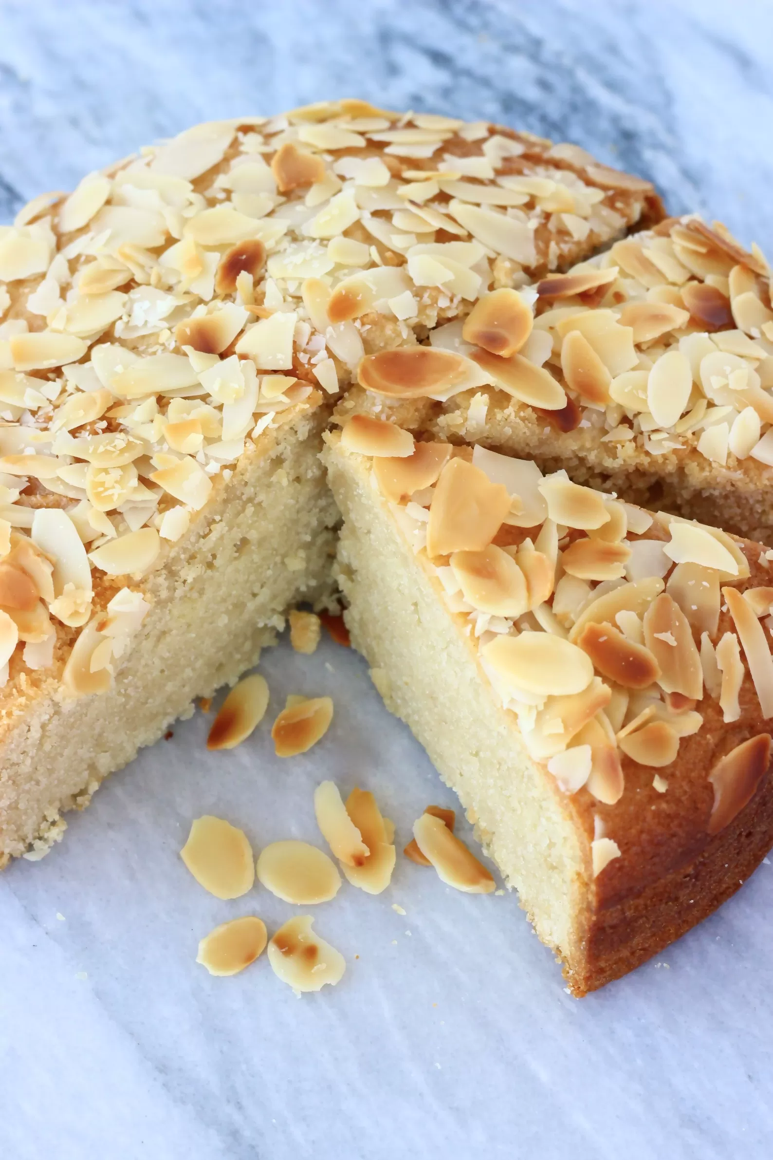 A round gluten-free vegan almond cake topped with flaked almonds cut into slices on a marble background