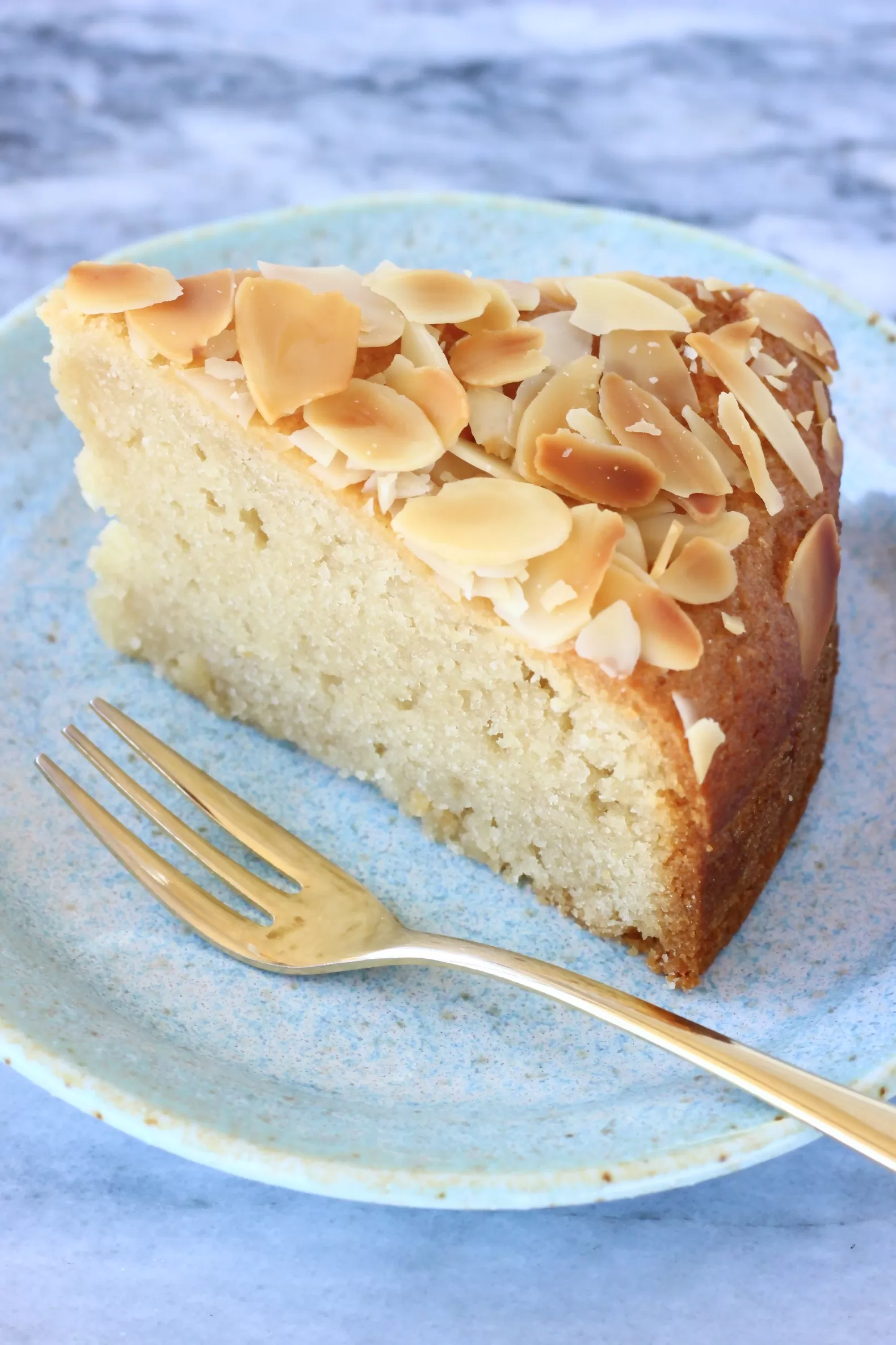 A slice of gluten-free vegan almond cake on a blue plate with a gold fork