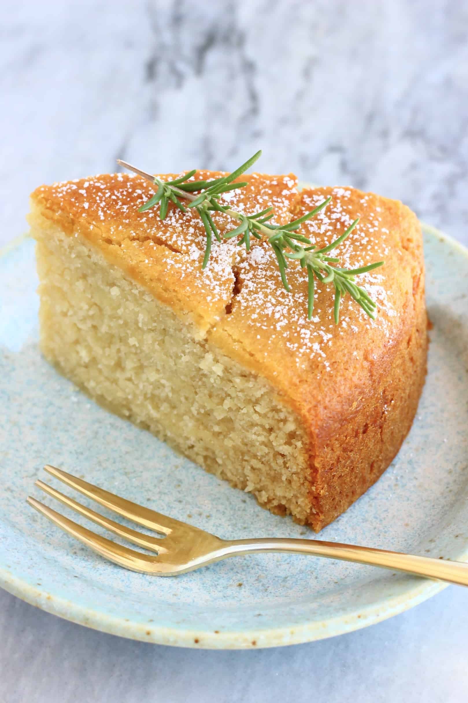 A slice of gluten-free vegan olive oil cake decorated with rosemary on a plate with a fork