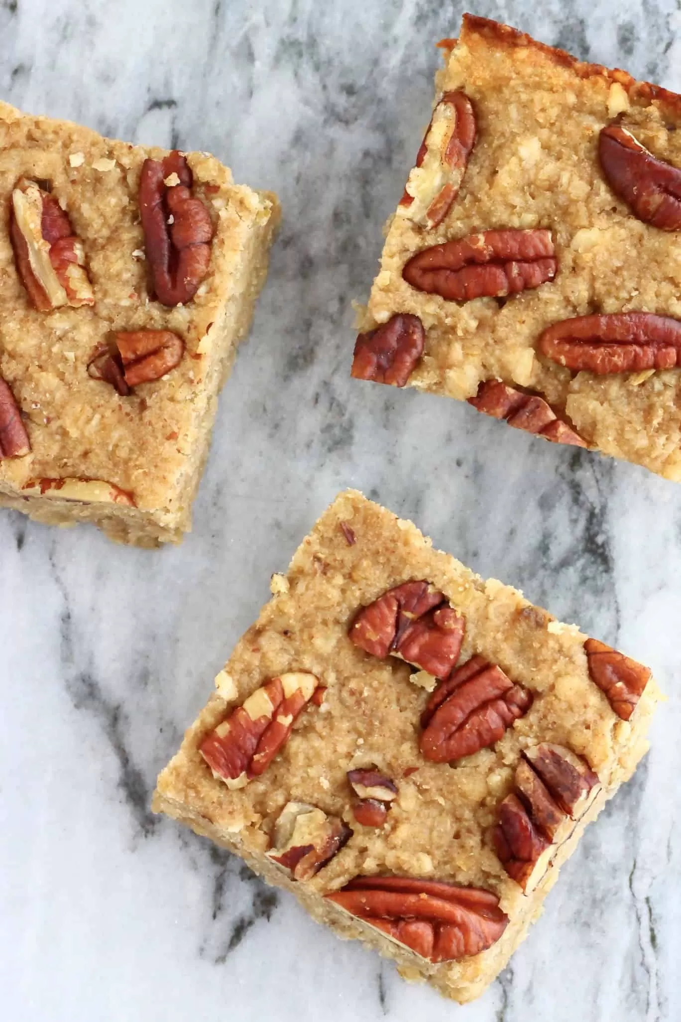Three square baked banana oatmeal bars topped with pecan nuts on a marble background