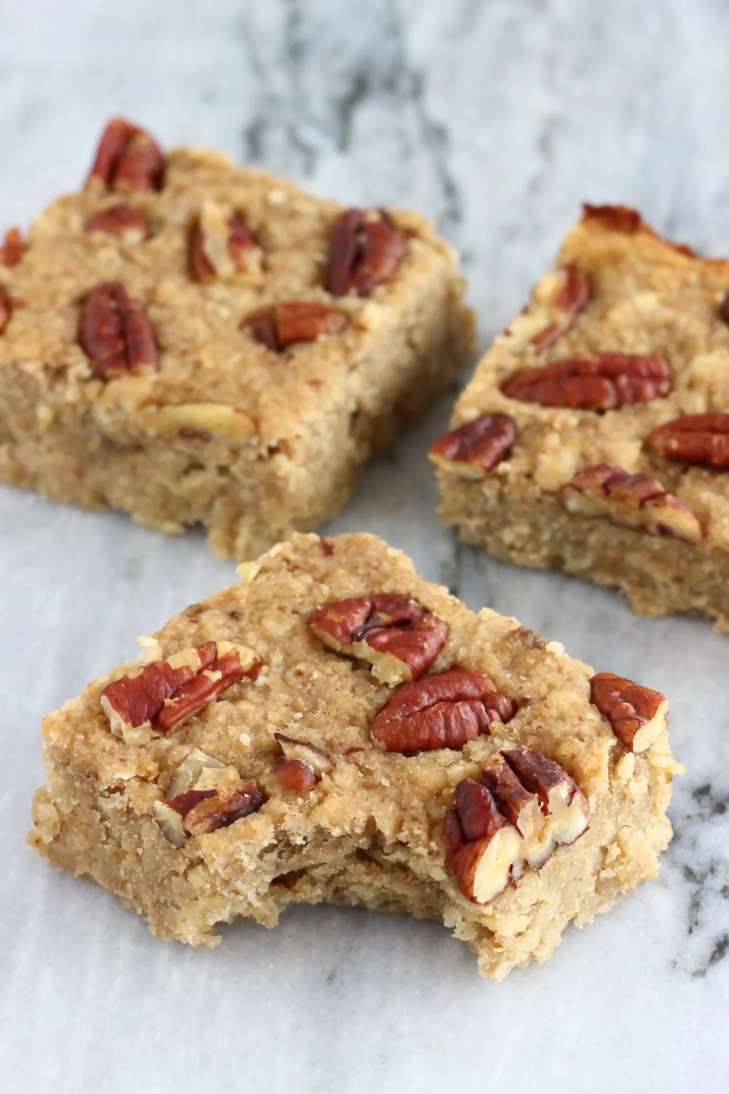 Three baked banana oatmeal bars topped with pecan nuts on a marble background, with a bite taken out of one
